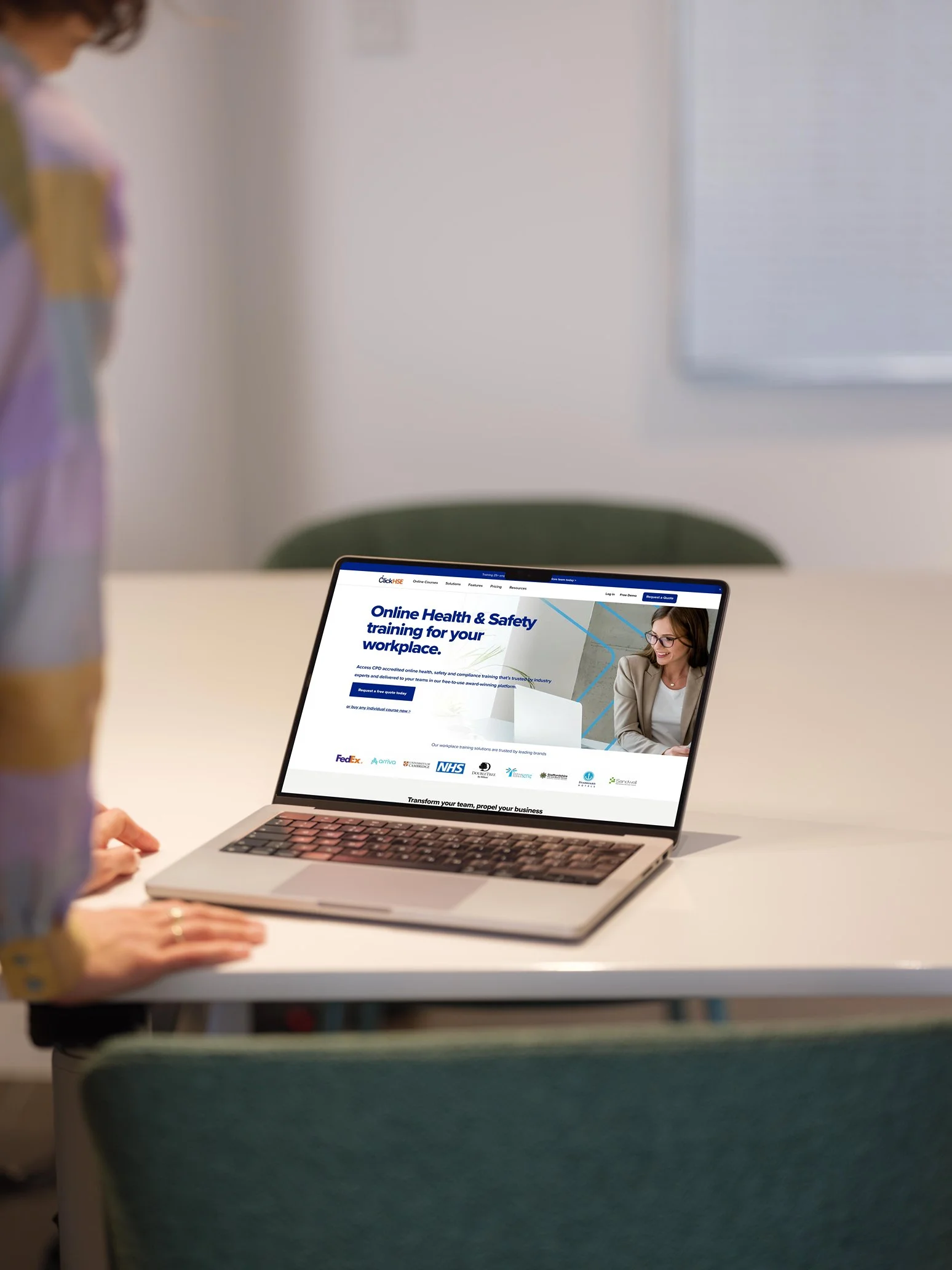 A laptop on a white table displays a webpage titled 'Online Health & Safety training for your workplace.' A person with a blurred face is seated nearby, and a vase with flowers is visible on the left side of the image.