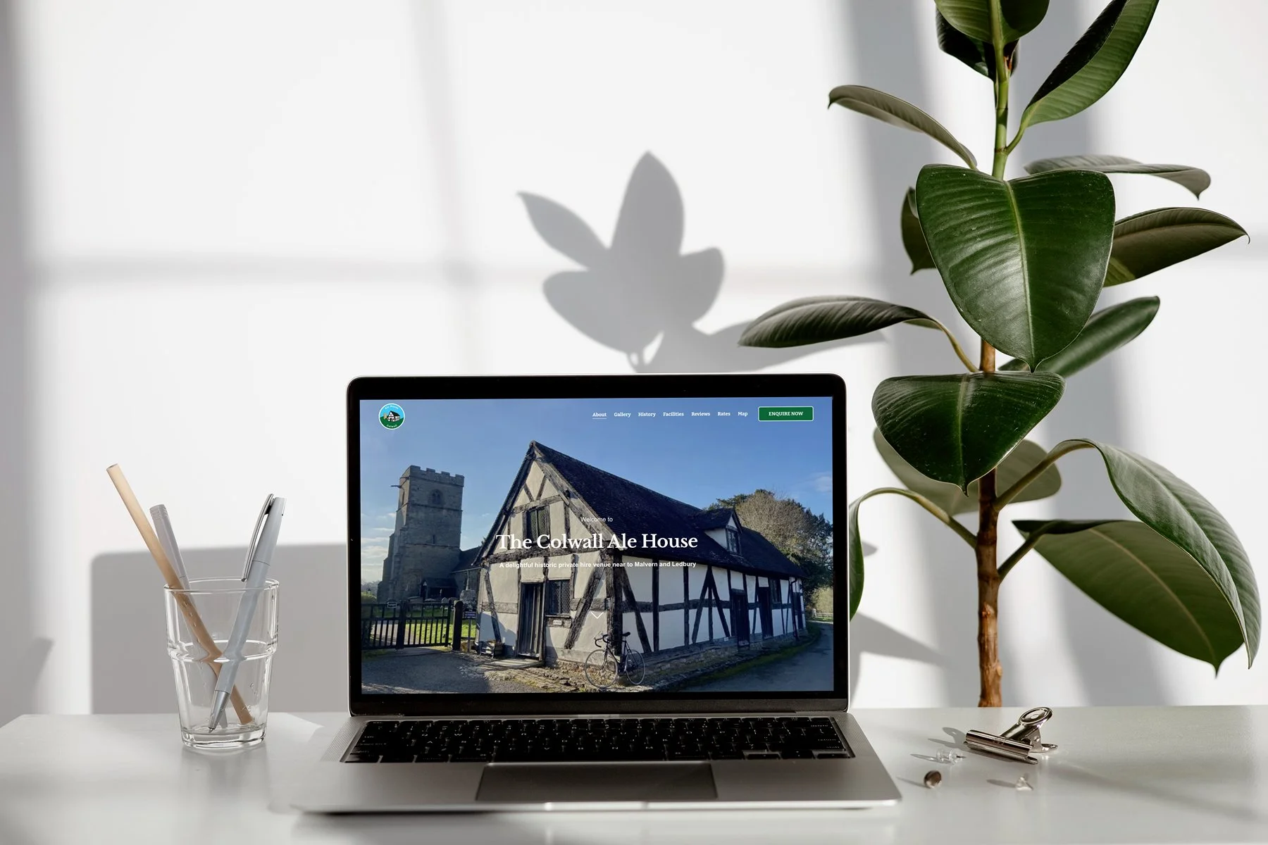 A laptop on a white desk displaying a website about The Coldwall Ale House, with a historic building and a bicycle outside. To the left of the laptop, there is a glass with three pens. On the right, there are binder clips and a paperclip. A large green leafy plant is behind the desk, casting a shadow on the wall.