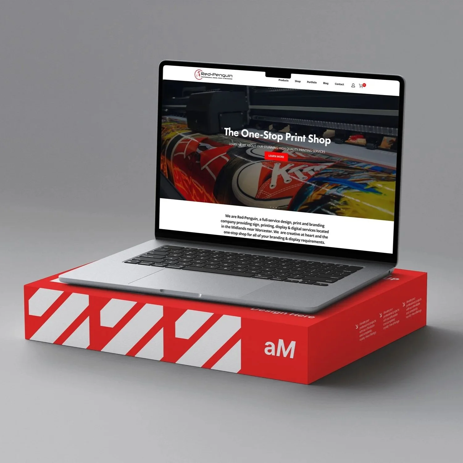 A laptop displaying a print shop website sits on top of a red and white striped box. The website has a header with navigation links and showcases high-quality printing services, featuring an image of a large format printer printing colorful graphics.