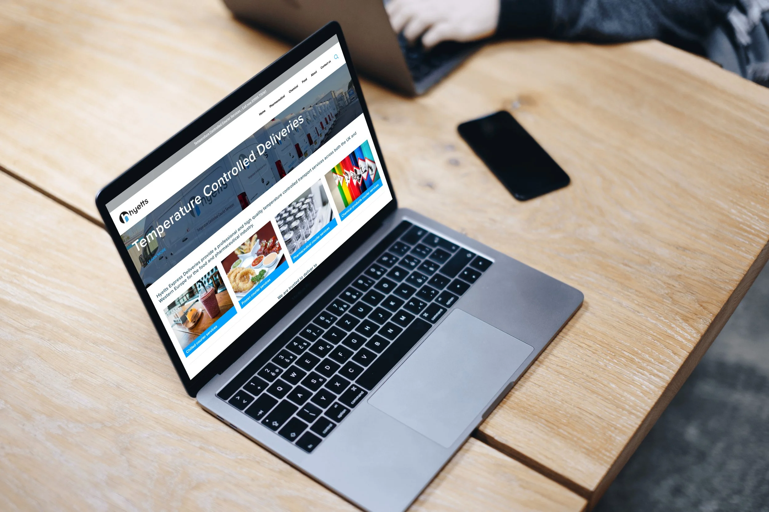 A silver laptop on a wooden table displaying a website titled "Temperature Controlled Deliveries" with images of trucks, food, desserts, and colorful graphics.