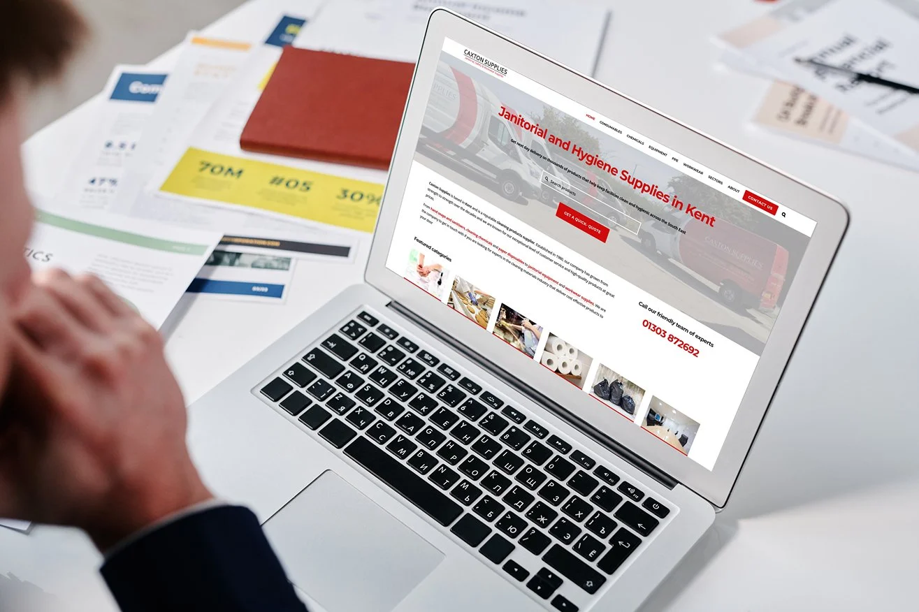 Person viewing a website on a laptop screen about janitorial and hygiene supplies in Kent, with various papers and brochures scattered on the white desk.