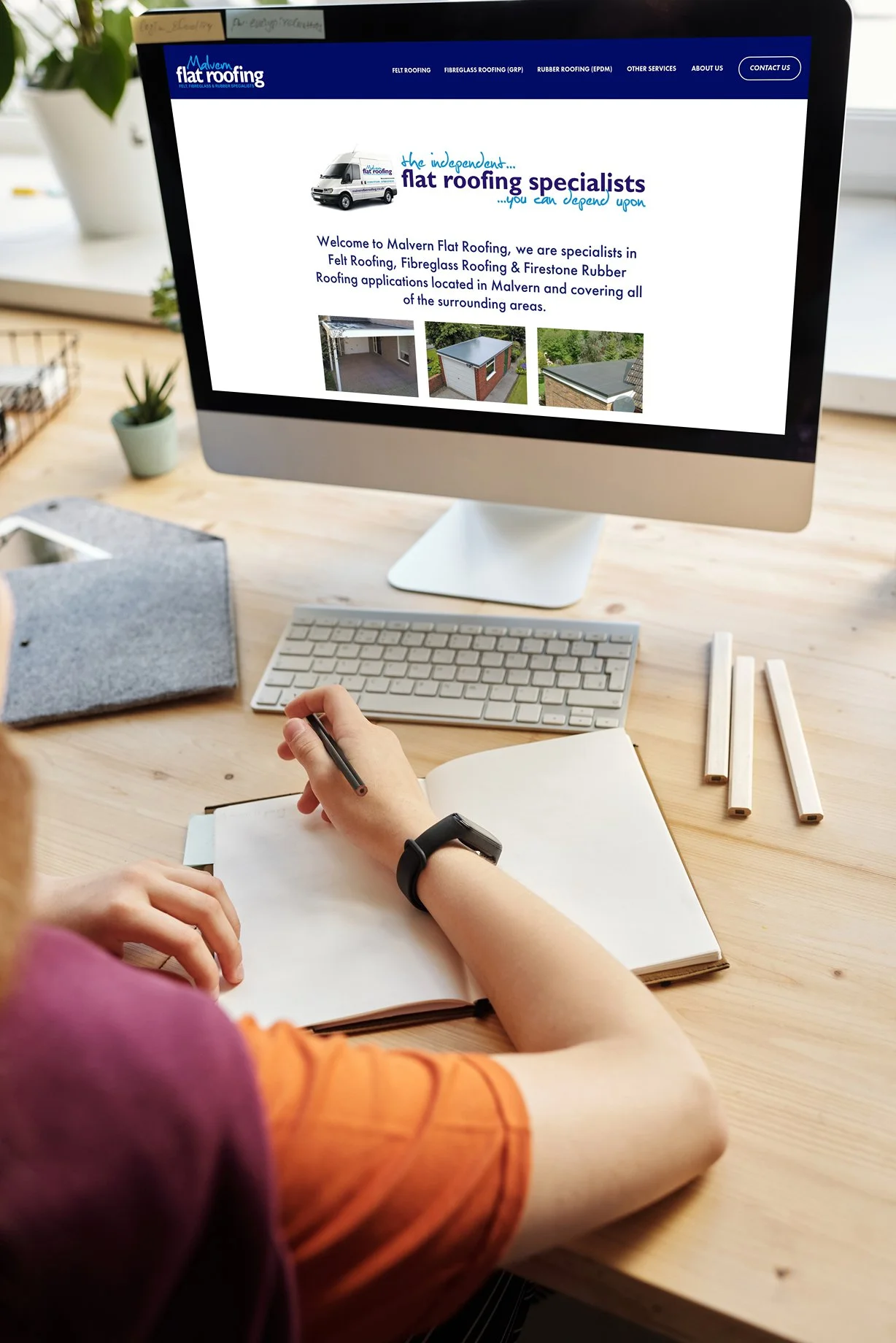 Person taking notes while working on a computer with a roofing company's website displayed on the screen, on a light wooden desk with a potted plant, a notebook, and wooden sticks nearby.