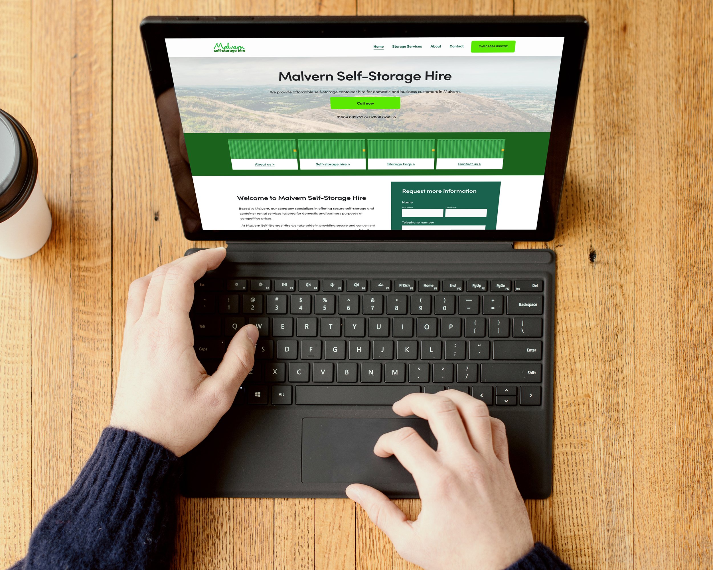 A person's hands using a computer keyboard on a wooden desk, with a tablet displaying a website for Malvern Self-Storage Hire. The website features a green and white theme with a landscape background and information about self-storage services.