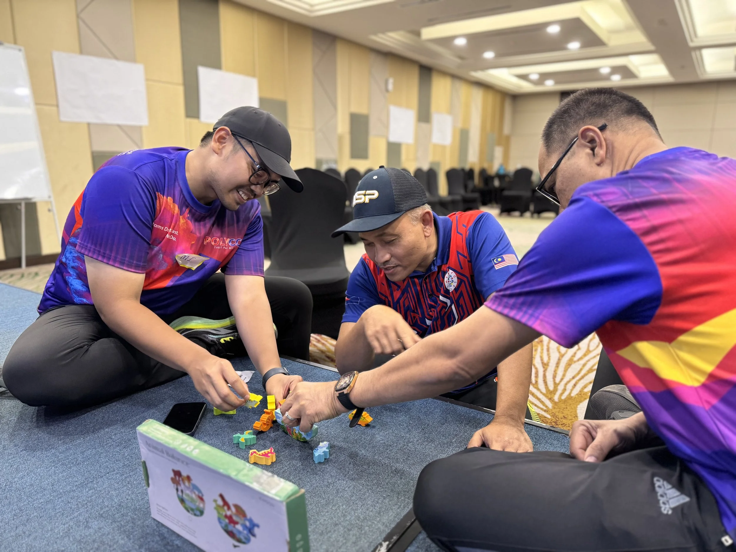 Three men wearing colorful shirts are playing a colorful board game on a table in a conference room. They are smiling and focused on the game pieces.