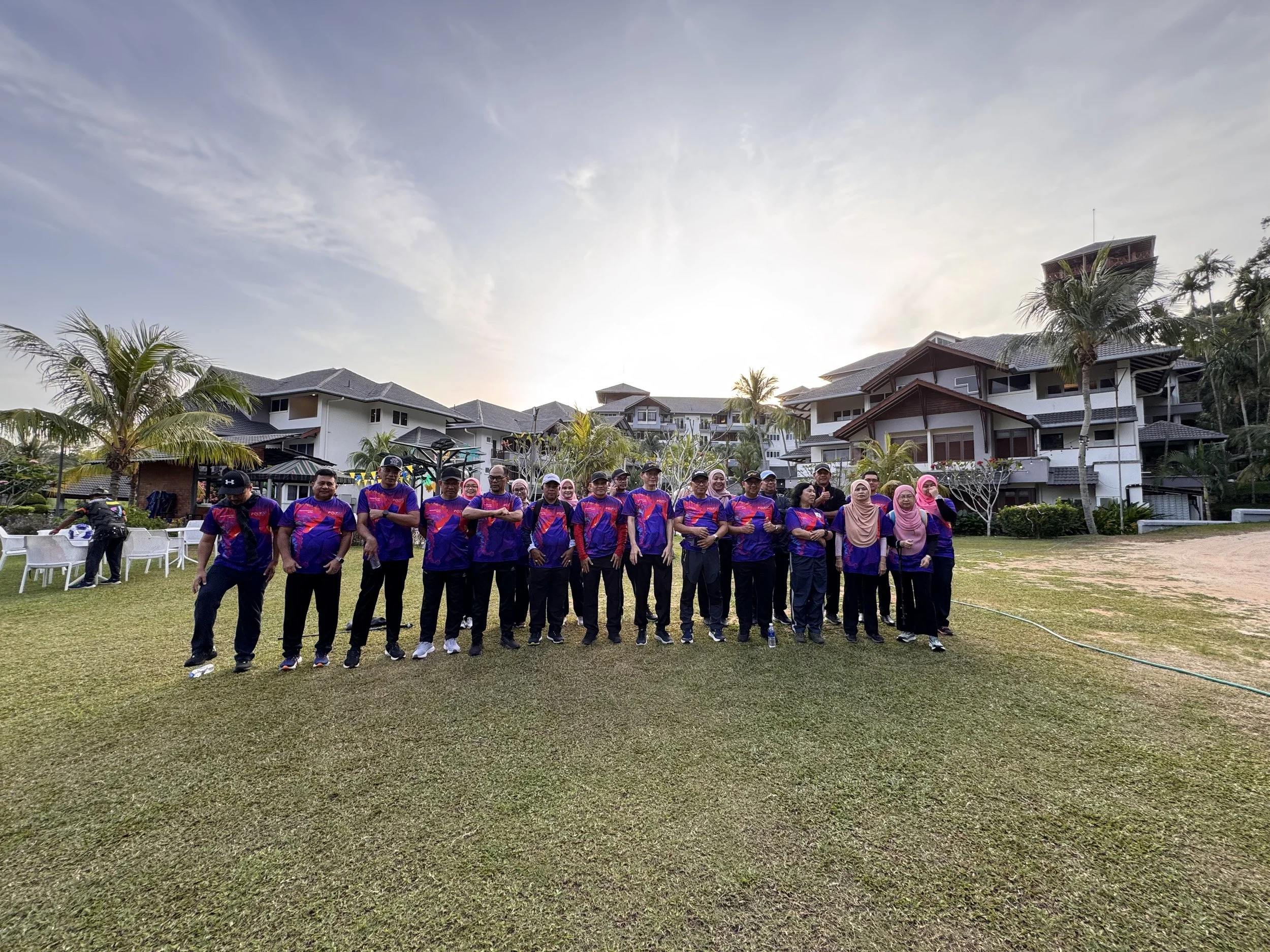 Group of people standing outdoors on grass field in front of large buildings, some palm trees, and cloudy sky during sunset.