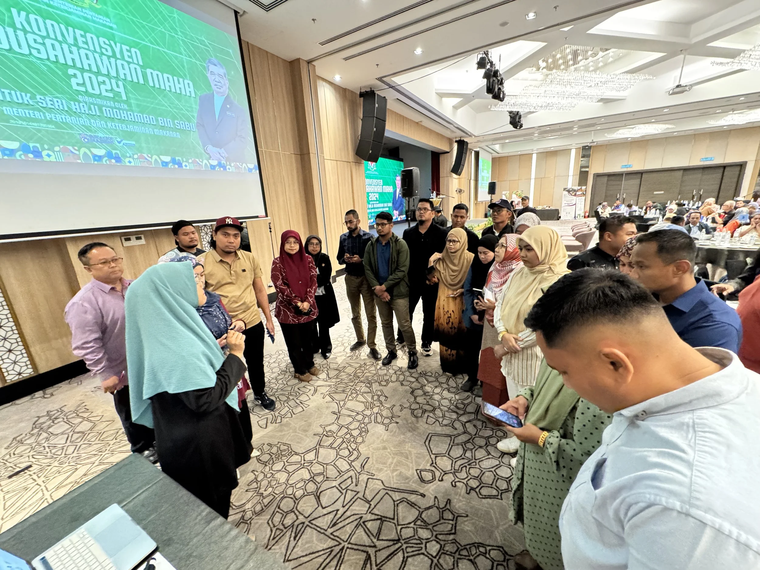 A group of people standing in a circle and paying attention to a woman speaking at the front of a conference room. The room features a large screen displaying a presentation, decorated with wood paneling and chandeliers, with tables and seated attend