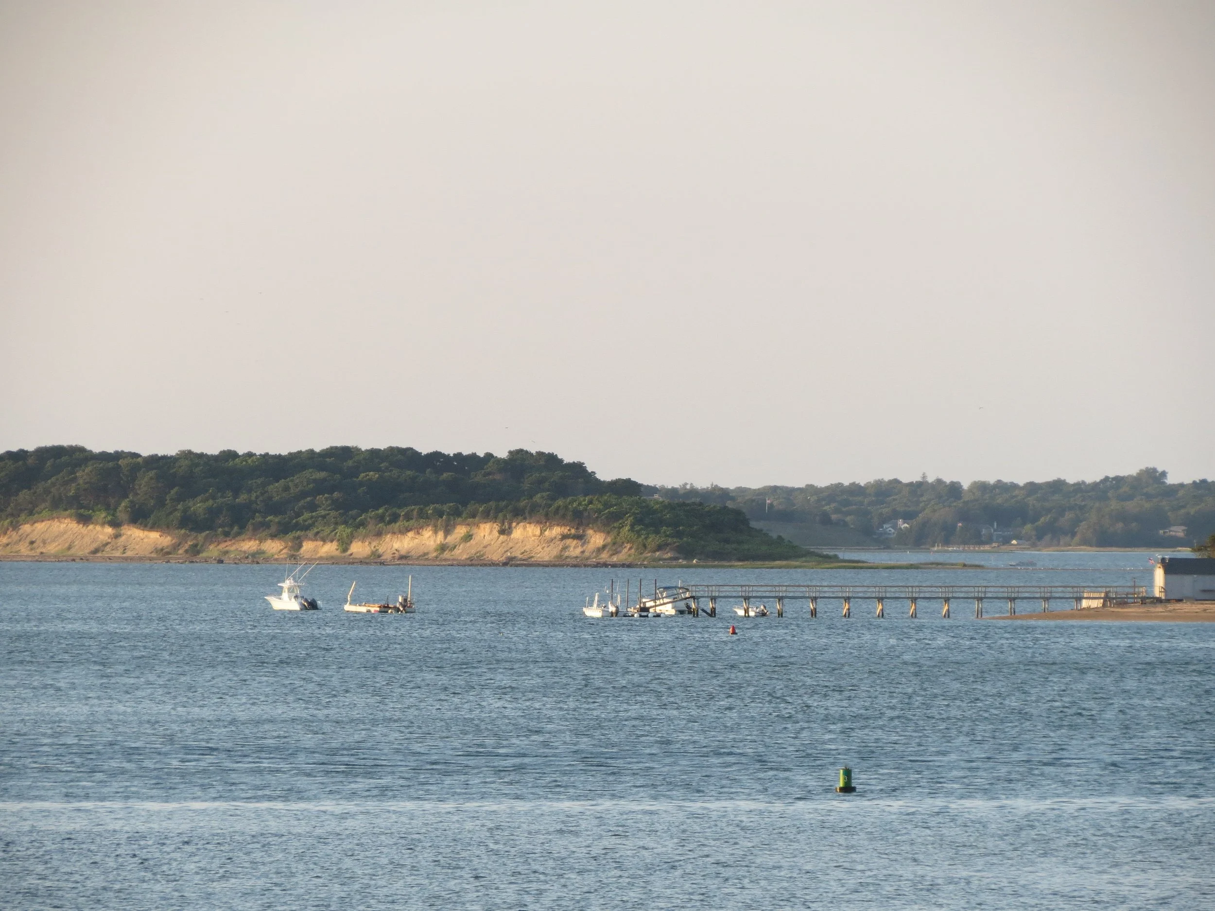 Calm water with boats near a pier, hillside with trees in the background, and a partly cloudy sky.