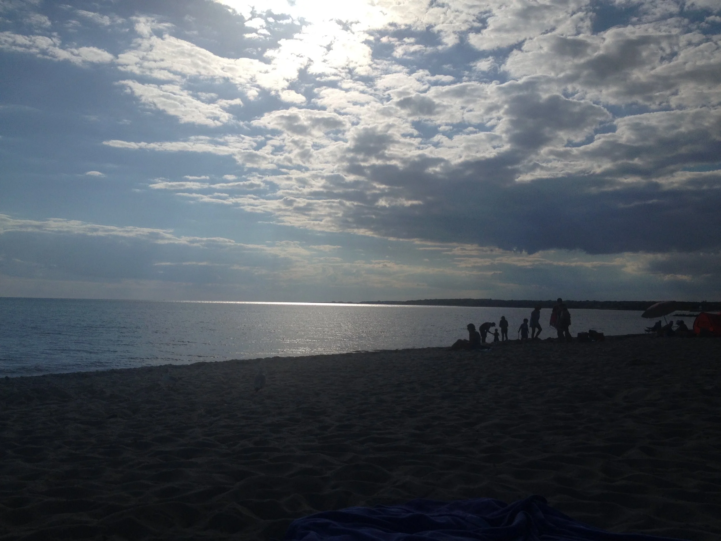 Beach scene during sunset or late afternoon, with a cloudy sky and the sun partially obscured. Silhouettes of people gathered on the sandy shore, some standing, bending, or sitting near a tent and umbrella.