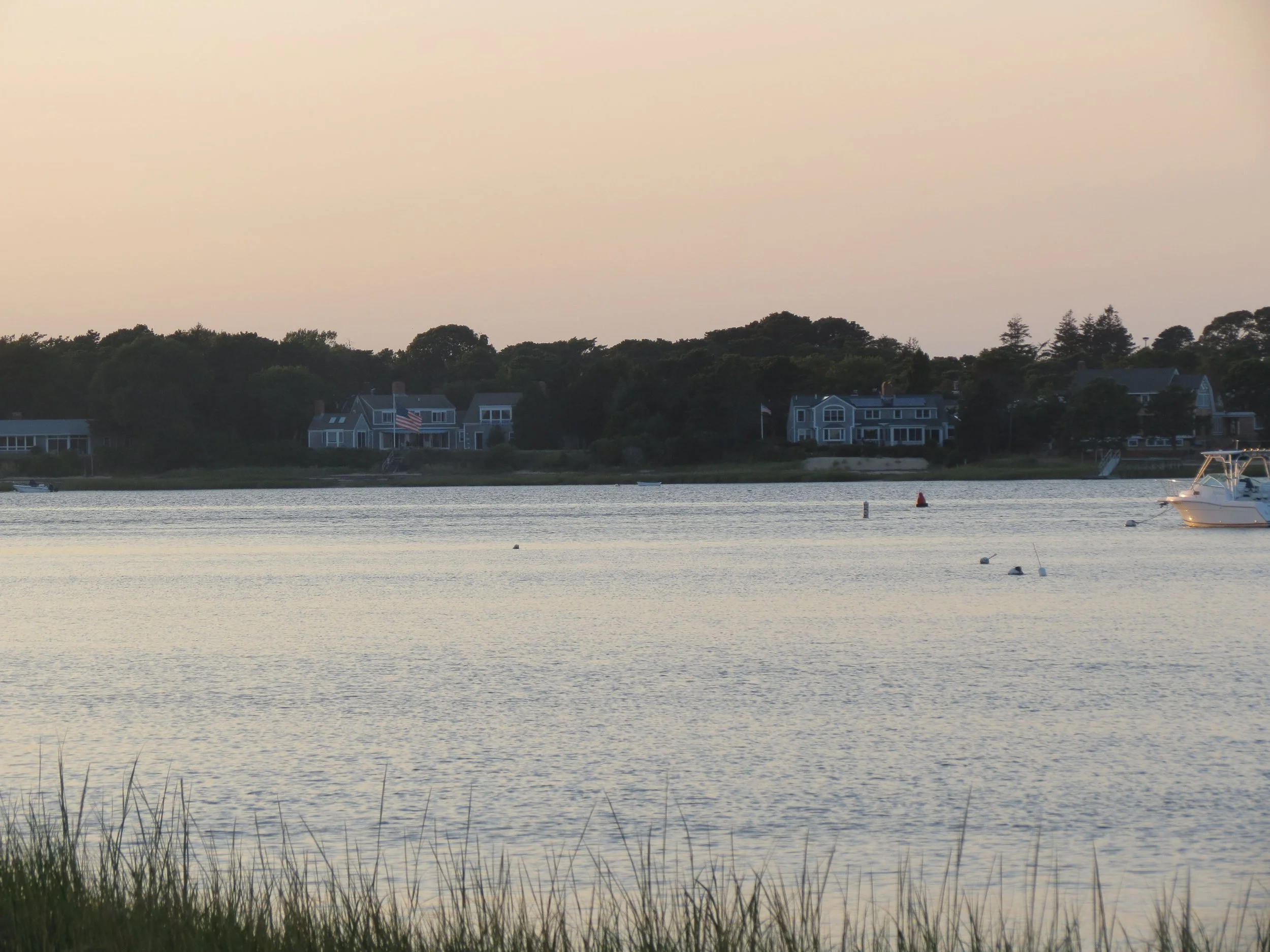 A peaceful waterfront scene at sunset with houses along the shoreline, some with American flags, and boats anchored in the water.