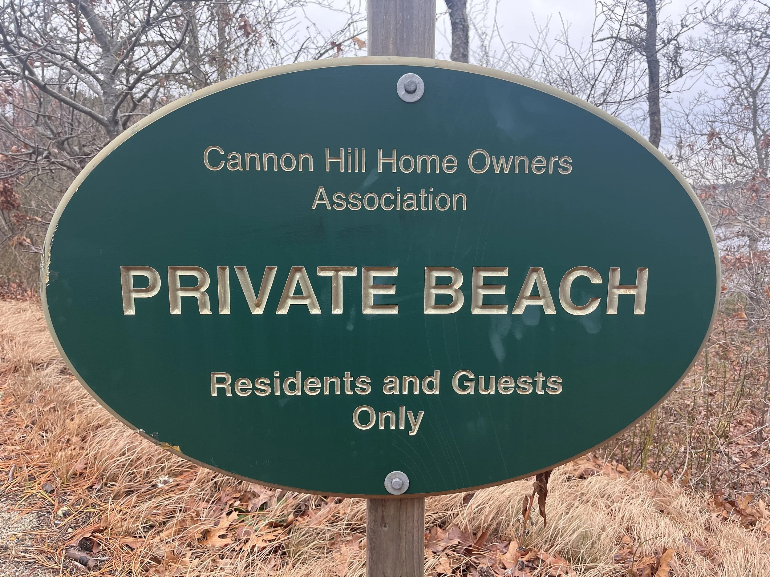 Green oval sign with silver text reading 'Cannon Hill Home Owners Association, Private Beach, Residents and Guests Only' in a wooded area with leafless trees and dry grass.