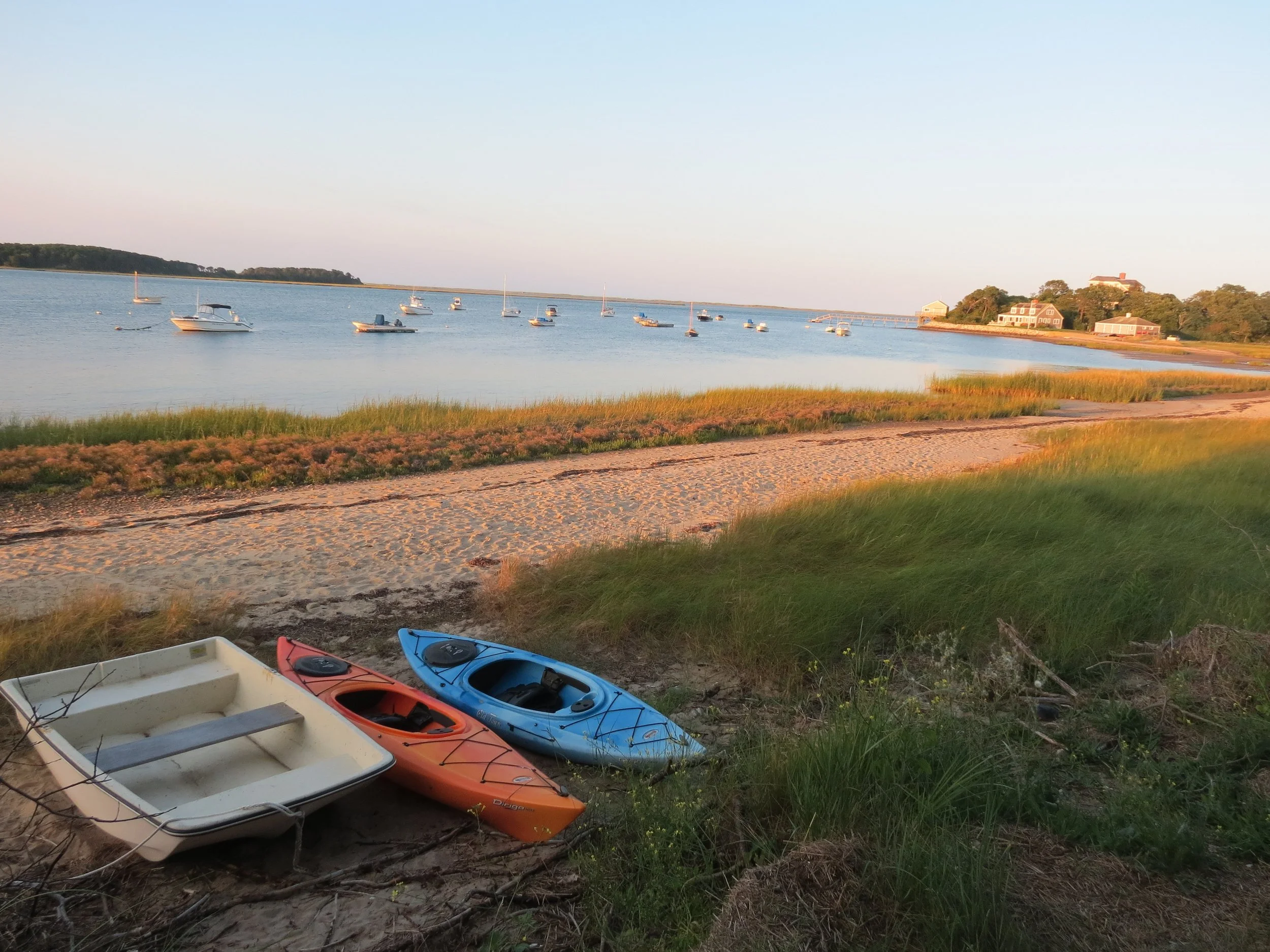 A beach scene during sunset with three kayaks and a small boat on the sand in the foreground, calm water with several boats anchored in the middle ground, and houses on a hill in the background.
