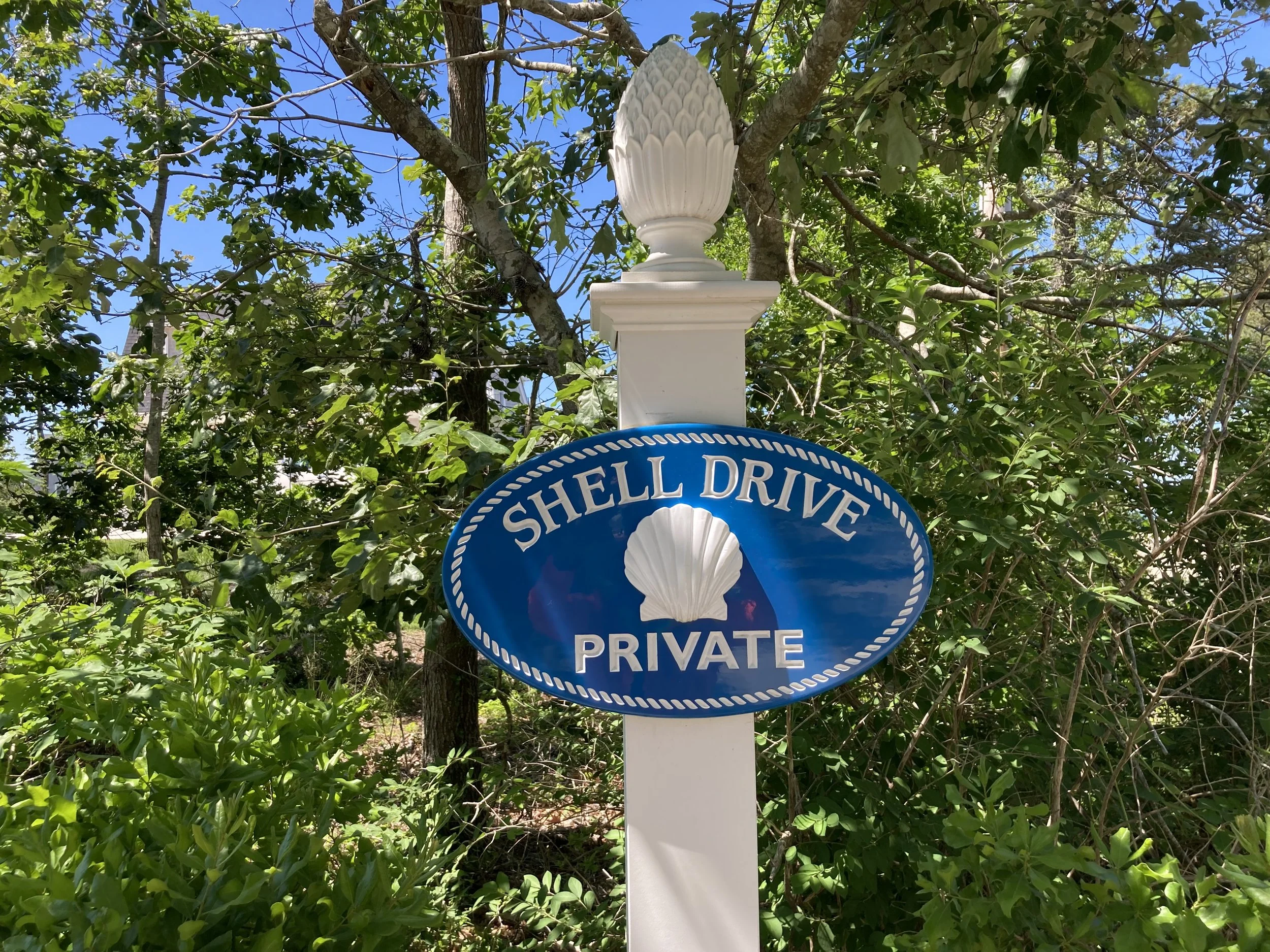 Street sign reading 'Shell Drive Private' with a shell symbol, mounted on a white post among green foliage and trees under a blue sky.