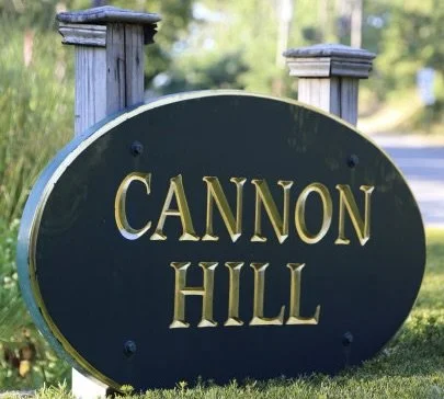A black oval sign with gold lettering that reads 'Cannon Hill,' mounted on two wooden posts on a grassy area, with trees and a road in the background.