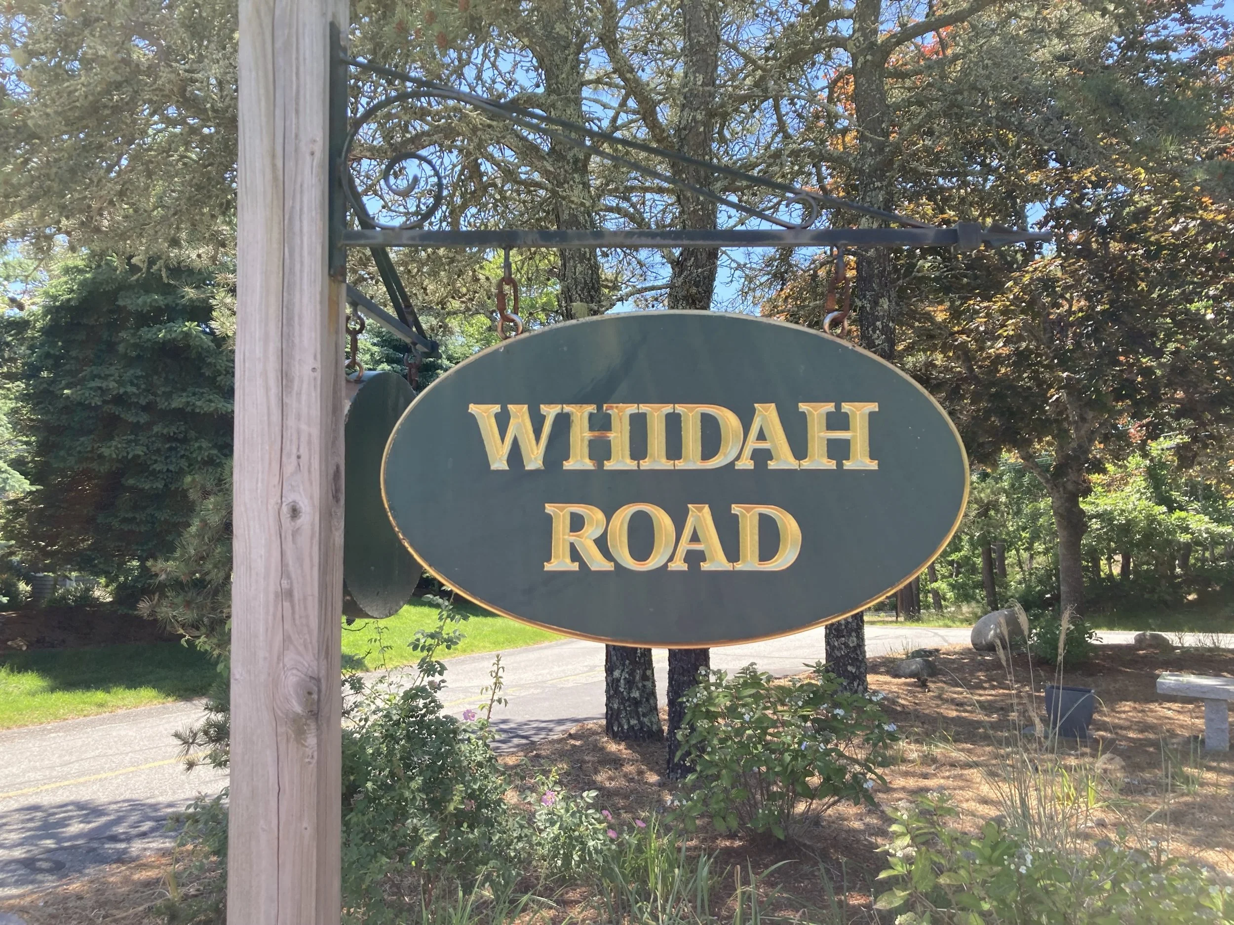 A green and yellow sign for Whidah Road hangs on a black metal frame, with trees, bushes, and sunlight in the background.