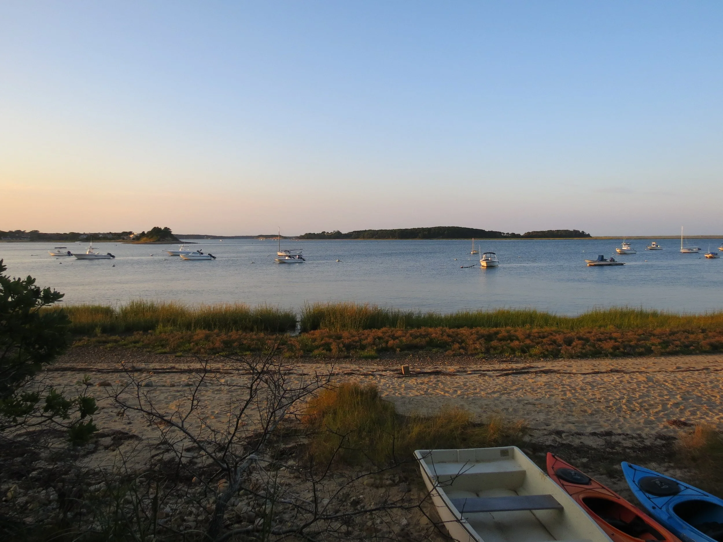 A serene lakeside scene at sunset with boats anchored in calm water and kayaks on sandy shore.