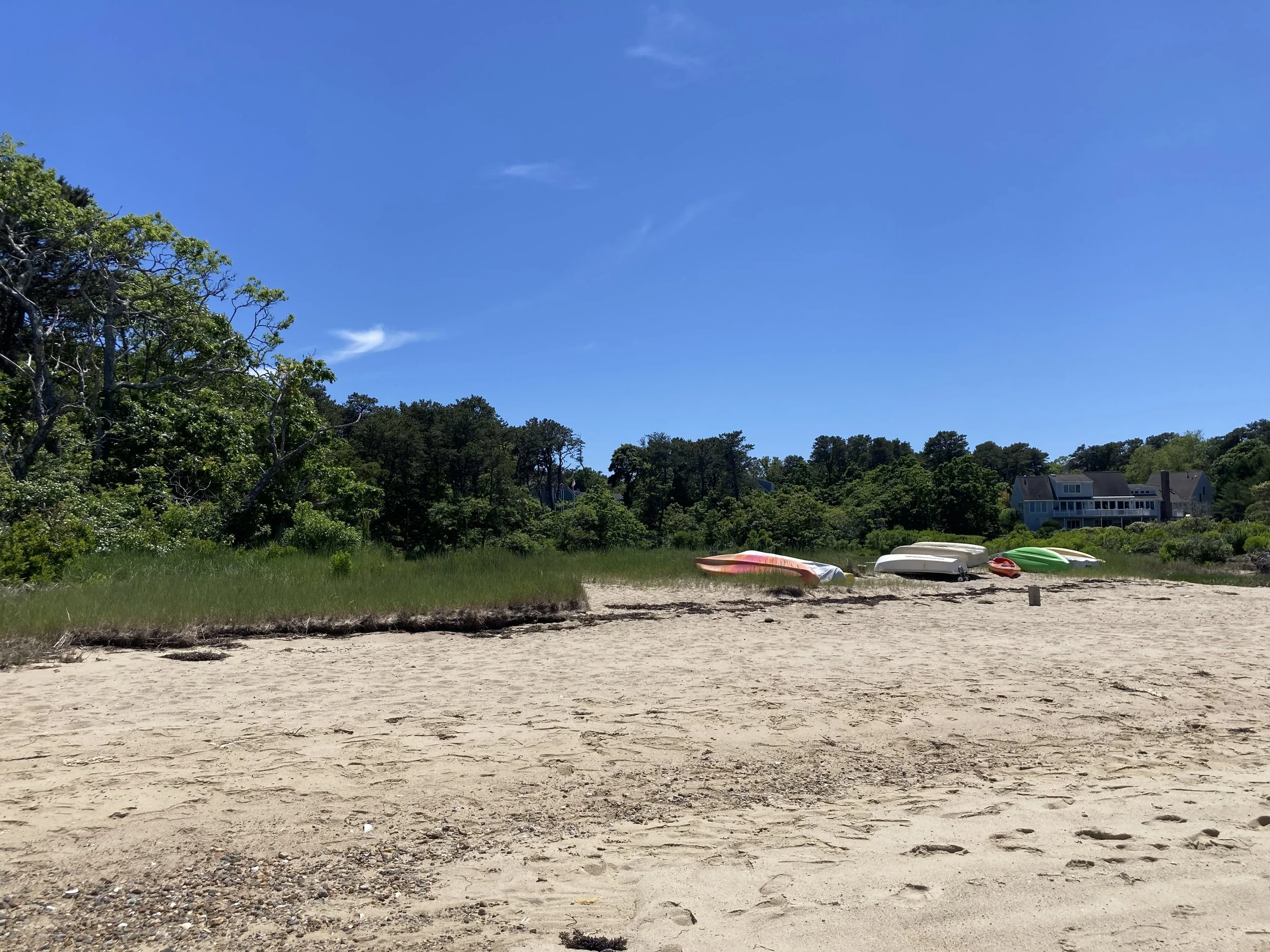 Beach with overturned boats, sandy shore, green trees, and a house in the background under a blue sky.