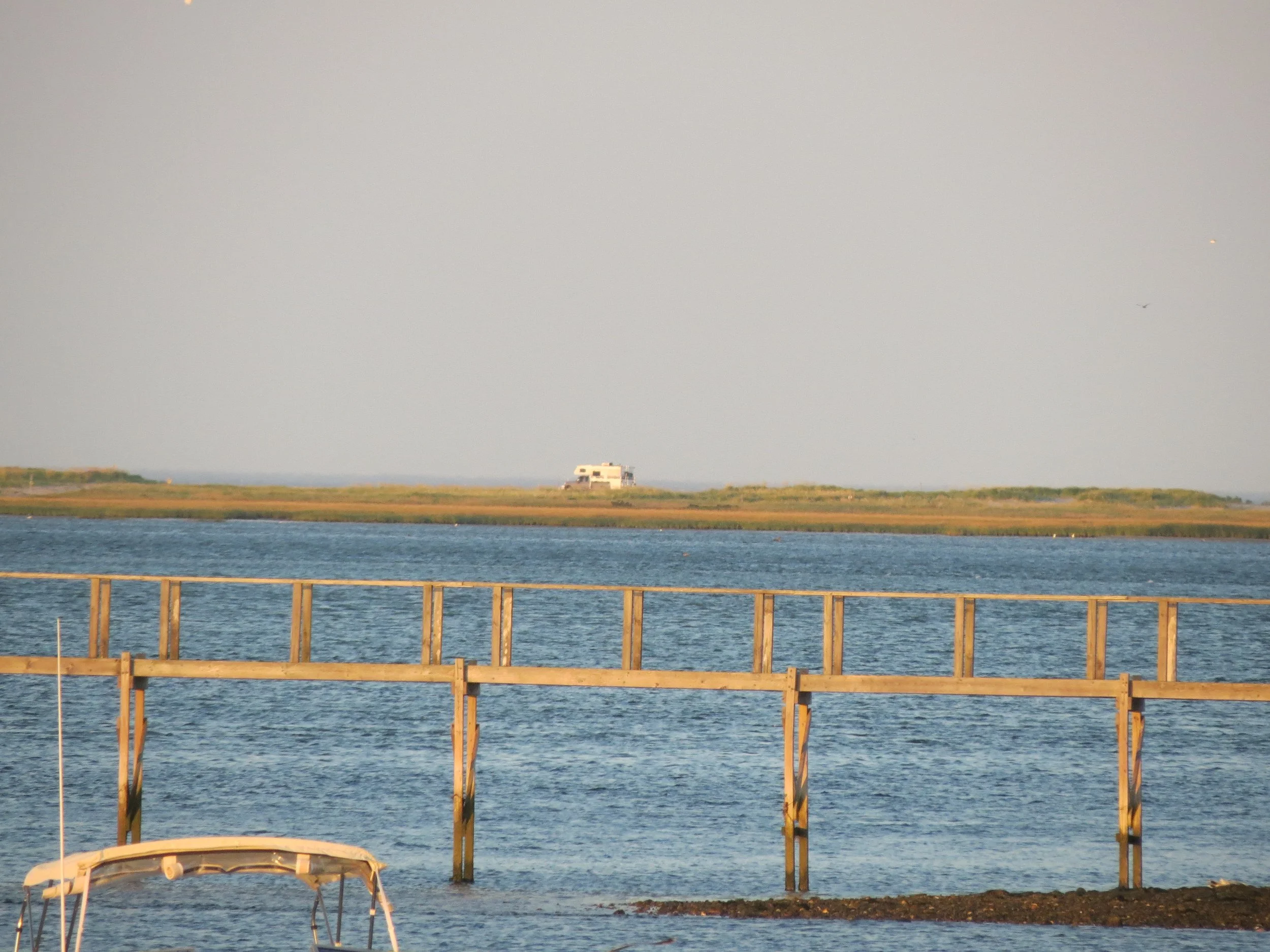 A boat on the water near a wooden dock, with a grassy shoreline and a white house or building in the distance under a clear sky.