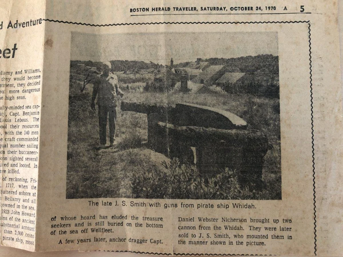 A black and white photo of a person standing on grassy land next to a small boat or submarine with a bridge and town in the background. The caption reads: "The late J. S. Smith with guns from pirate ship Whidah."
