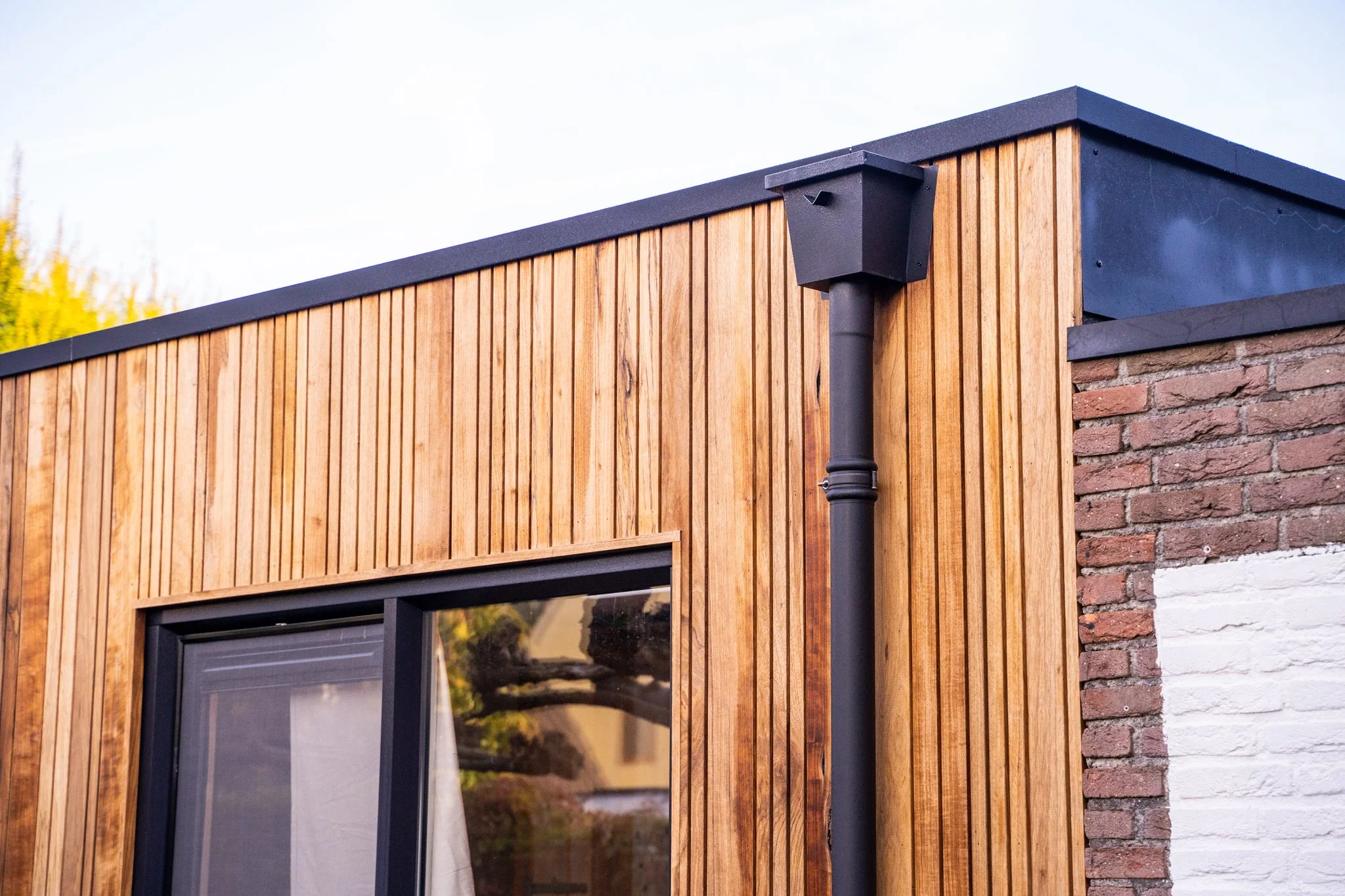 Modern huis met houten gevels en een zwarte regenpijp onder een dwarsgekante dakrand, met een grote raam en een deel van een witte en bakstenen muur.