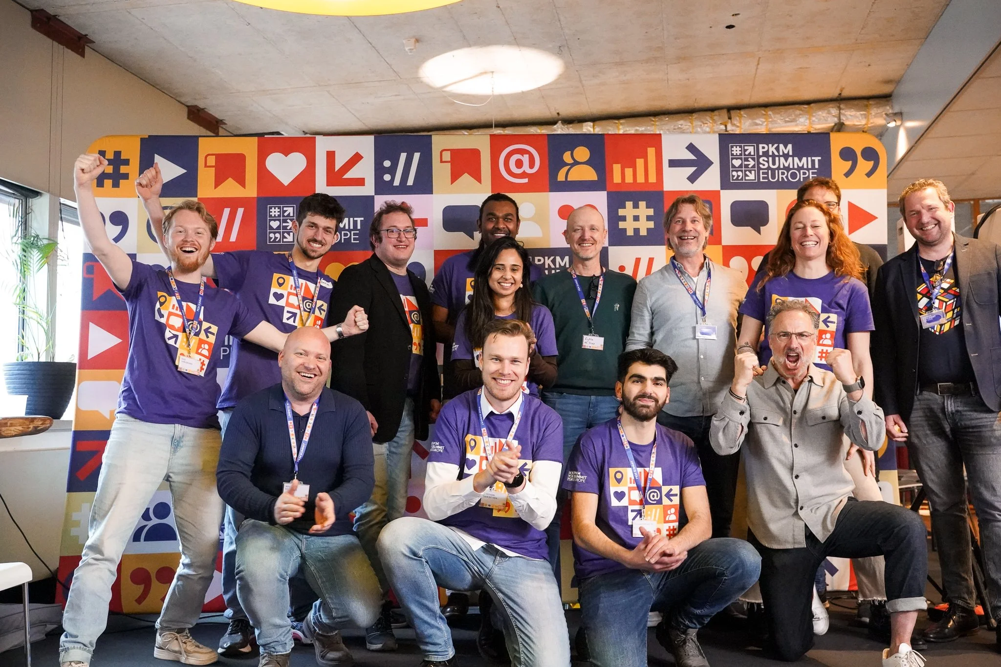 Een groep lachende mensen poseert voor een kleurrijke achtergrond met diverse iconen en het logo 'PKM Summit Europe'.
