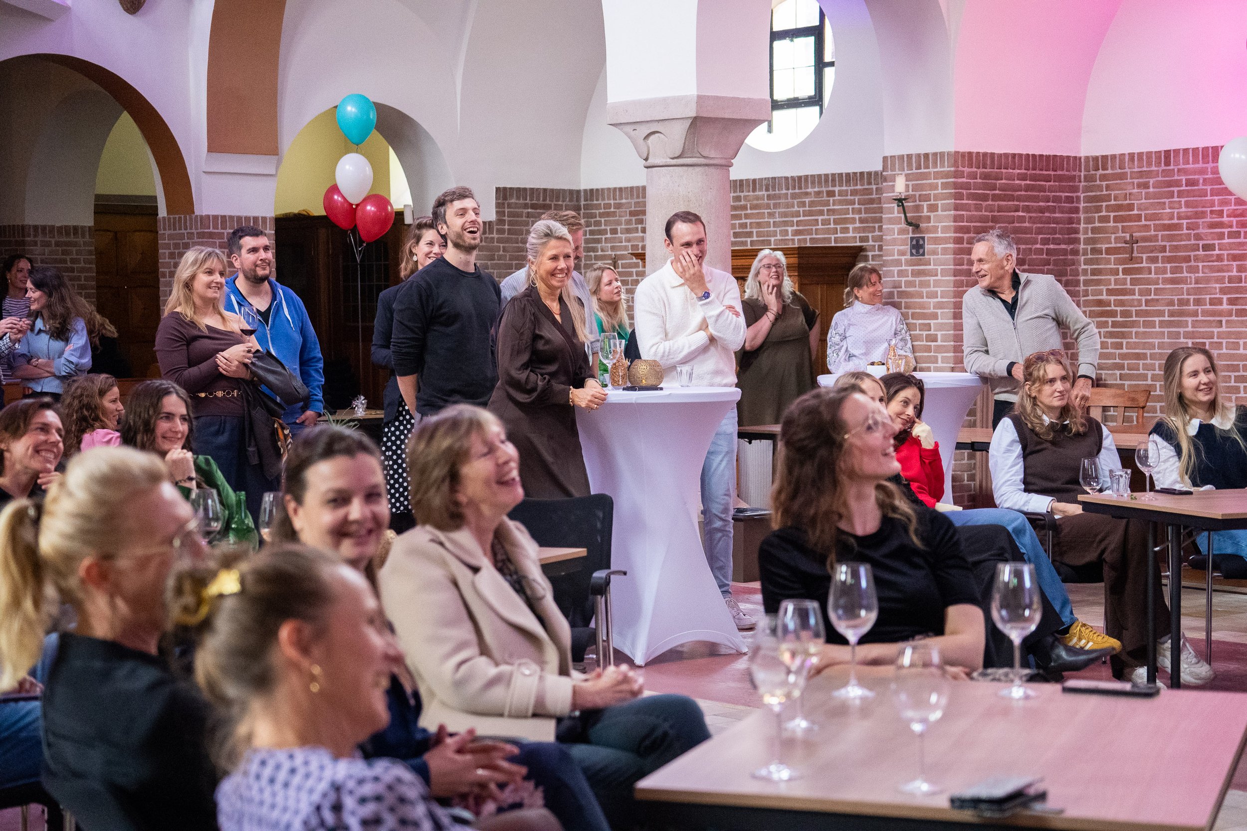 Een groep mensen lacht en kijkt naar een voorwerp of persoon, vermoedelijk tijdens een sociale bijeenkomst of feestje in een kerk of hall met gebogen plafonds en bakstenen muren.
