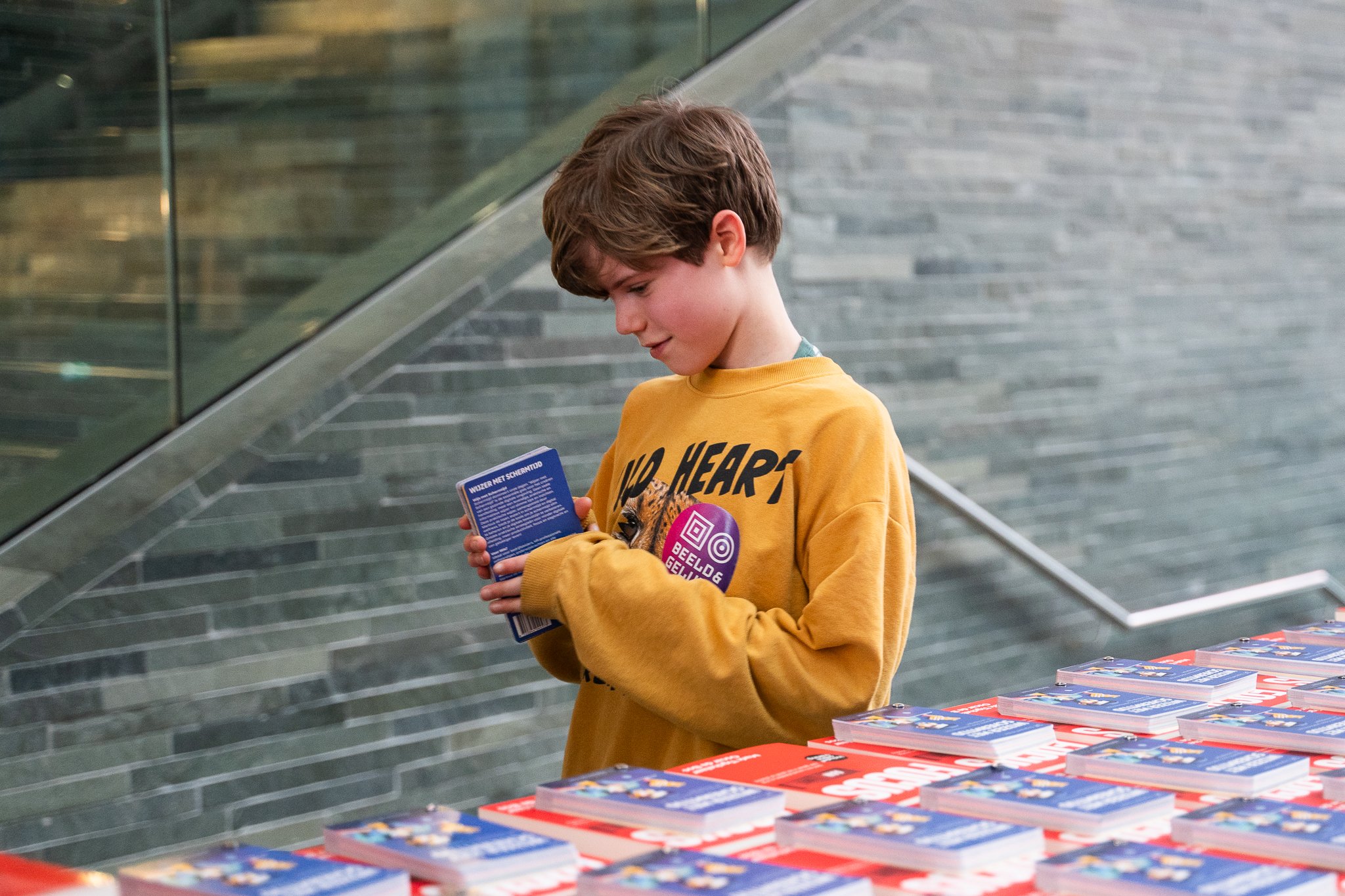 Jongen met bruin haar in een gele trui leest een boekje bij een tafel met meer boeken, binnen in een gebouw met een glazen trap en een muur van grijze stenen.