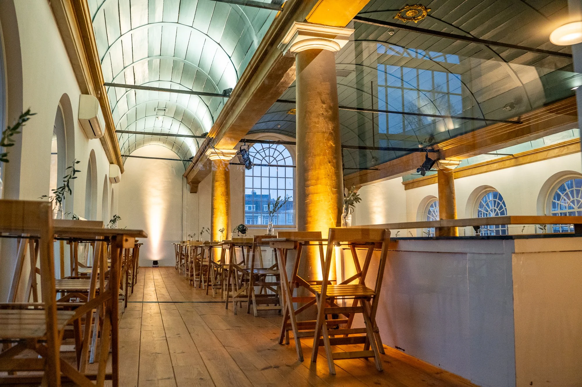 Binnenruimte met lange rij houten tafels en stoelen, grote glasramen, gouden kolommen, houten vloer, zachte verlichting en een glazen plafond met metalen balken.