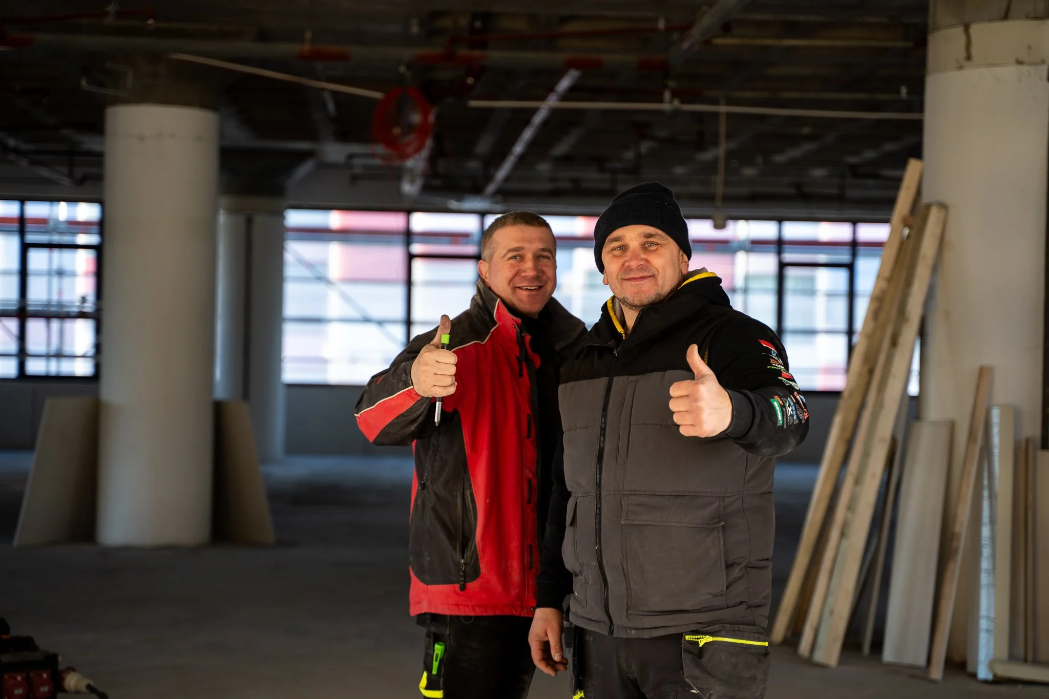 Twee mannen in bouwkleding staan binnen een gebouw onder constructie, lachen en geven duim omhoog.