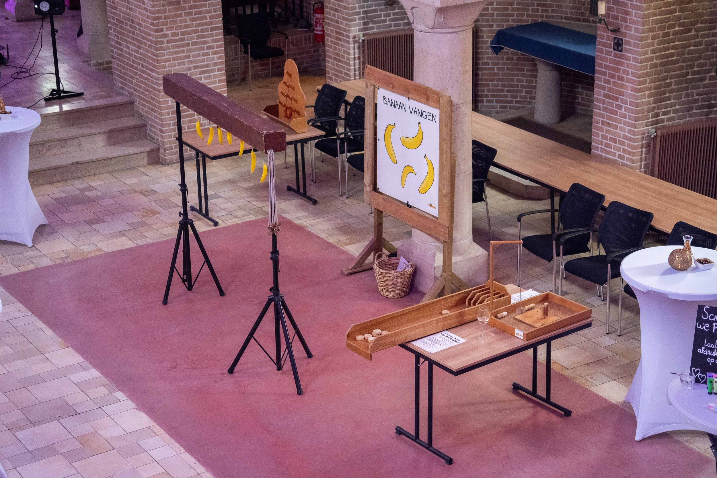 Binnenzalen met rood tapijt en tafels voor een spelactiviteit, waaronder een houten banenbaan voor het vangen van bananen, met meerdere bananenillustraties op een bord met tekst 'Banan vangen'.