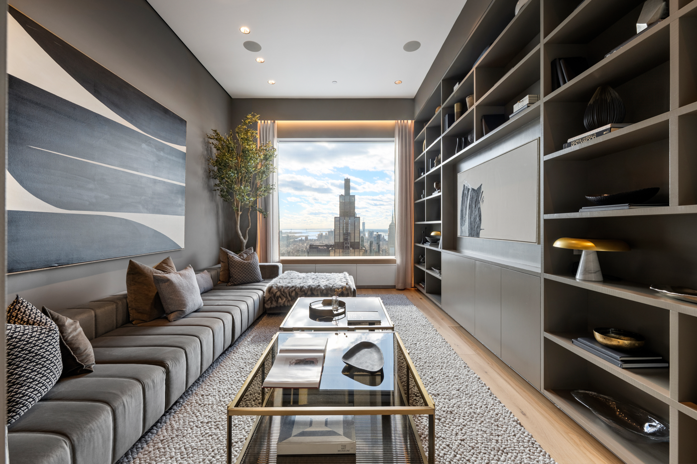 Modern living room with a large window view of a city skyline, featuring a long gray sofa with pillows, a glass coffee table, a textured area rug, and a tall shelf with decorative items.