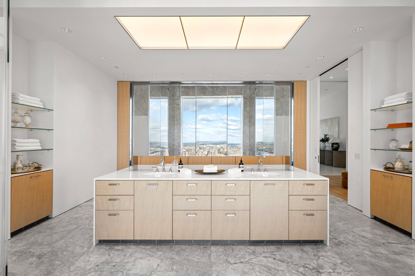 Modern bathroom with a large central vanity with two sinks, mirrors, and a city view through large windows.