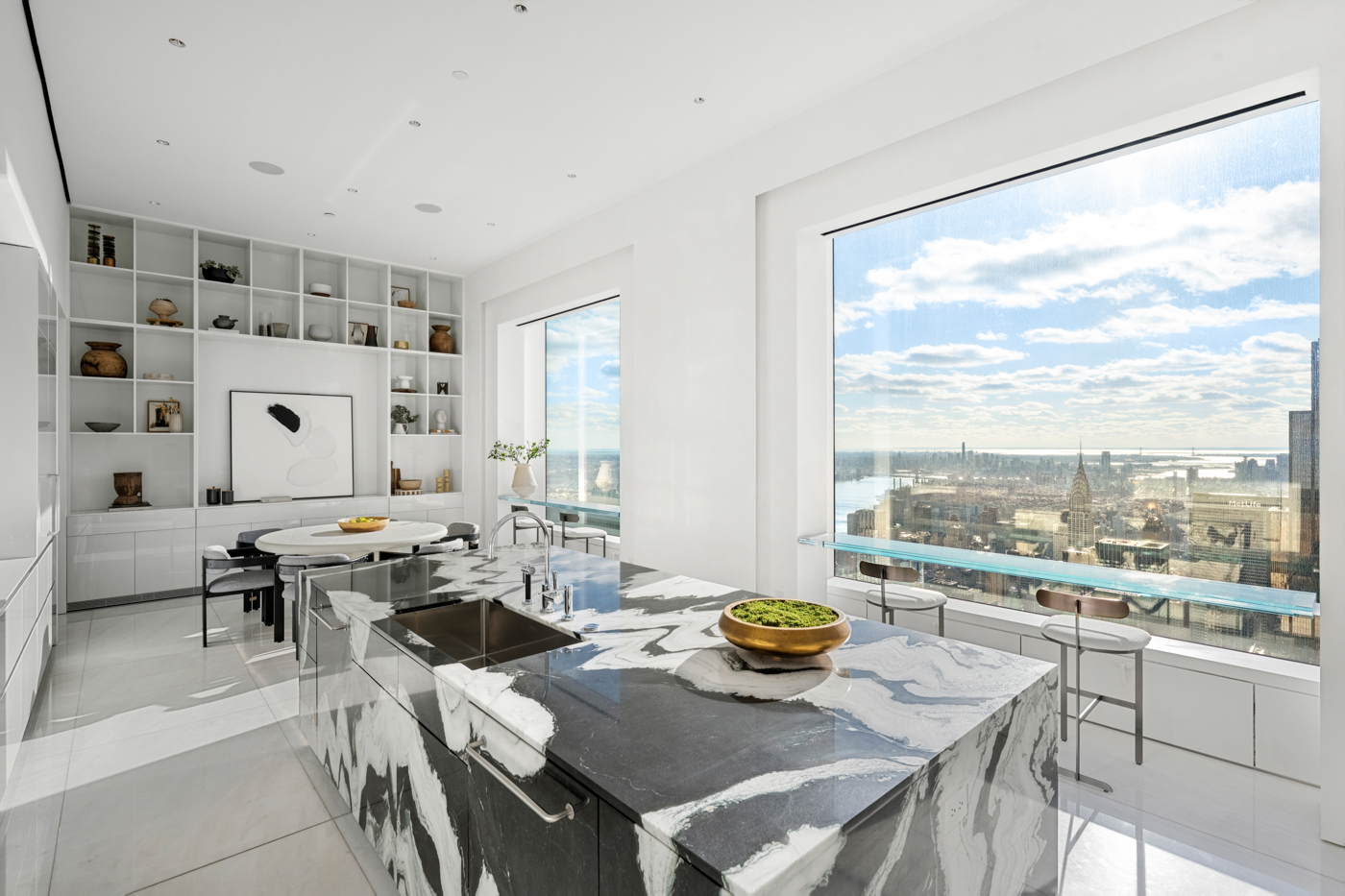 Modern kitchen with a black and white marble island, large windows with city view, and white shelving with decorative items.