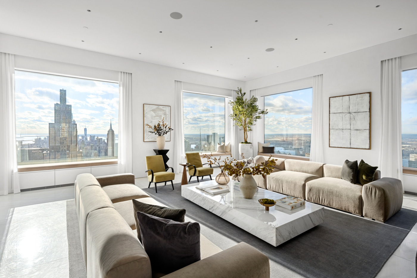 Modern living room with large windows overlooking a city skyline, featuring white walls, beige sofas, green armchairs, a marble coffee table with vases and books, artwork on the walls, and indoor plants.