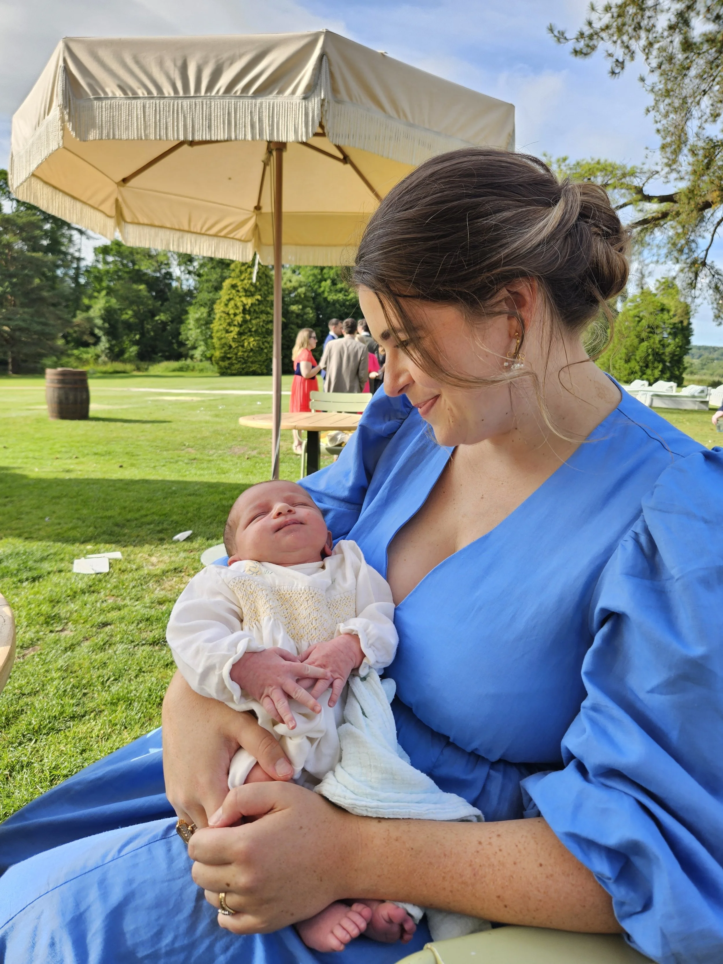 A woman in a blue dress holding a newborn baby outside under a cream-colored umbrella at a social gathering on a grassy lawn with trees in the background.