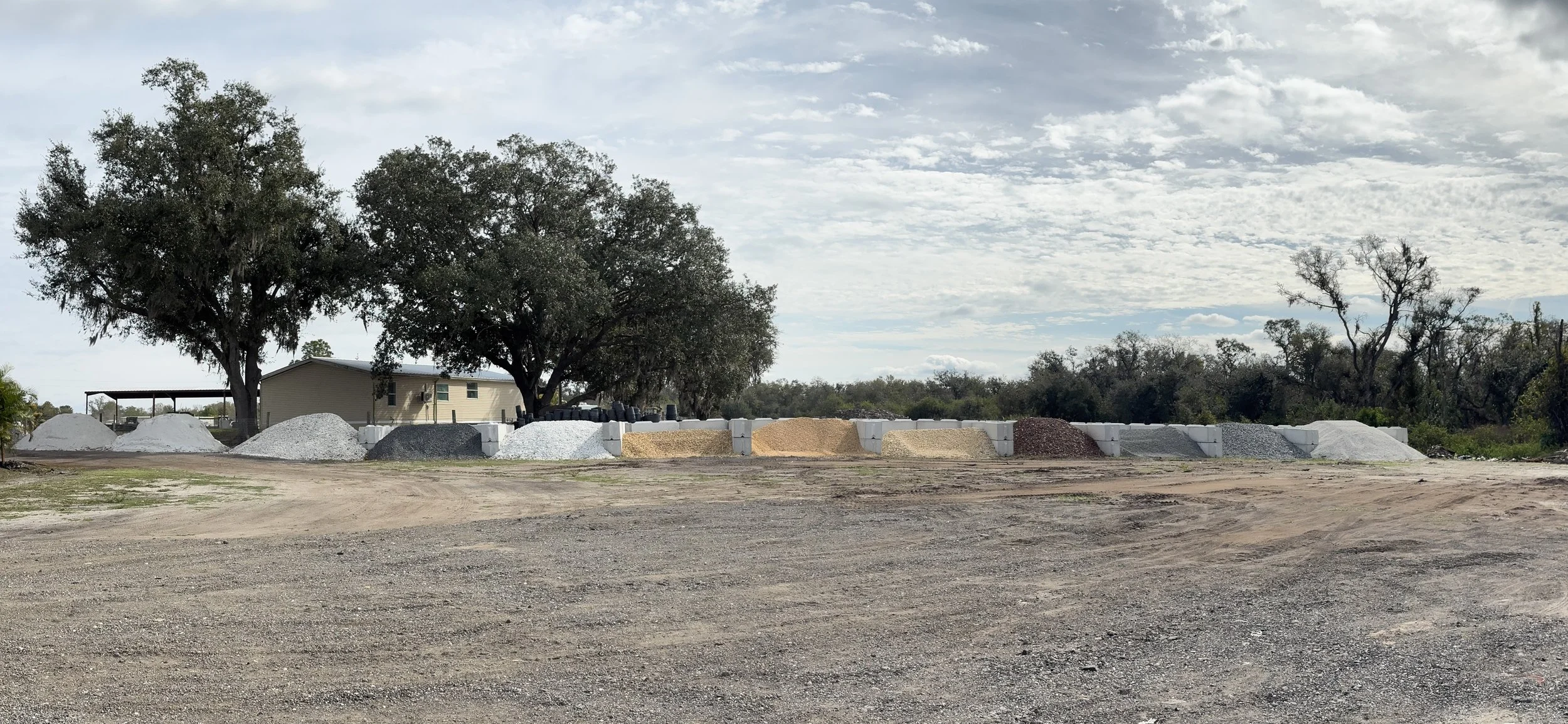 Various piles of gravel and sand aligned outdoors, with a house and trees in the background under a cloudy sky.