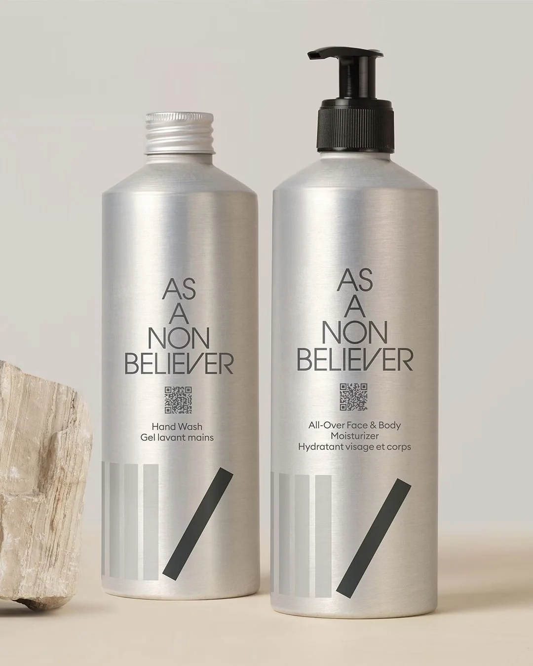 Two aluminium bottles, one with a screw cap and the other with a pump dispenser, labeled "As A Non Believer"; one bottle is hand wash, the other is all-over face and body moisturizer, both with QR codes and minimalist design on a neutral background.