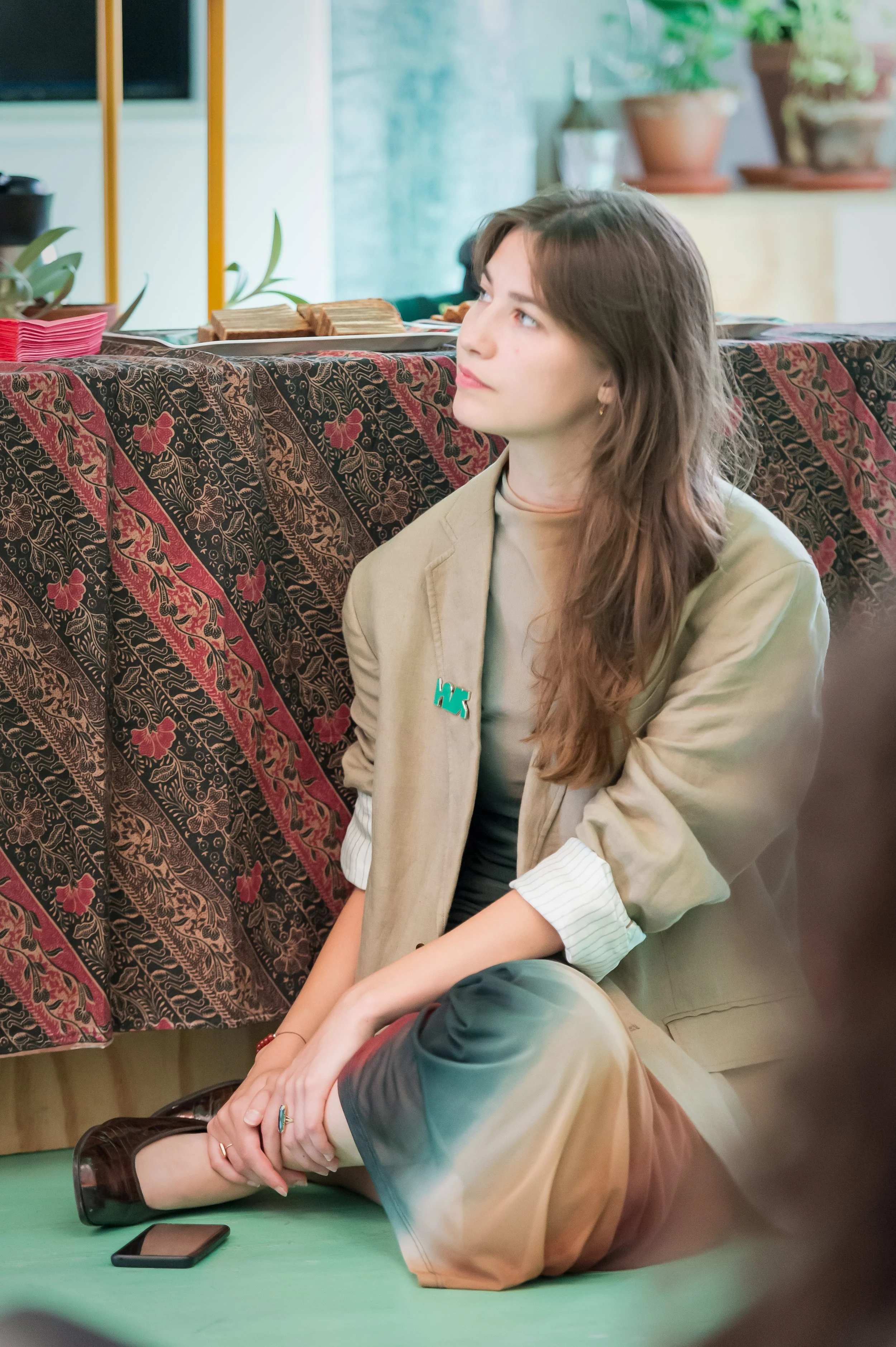 A young woman sitting on the floor with her legs crossed, wearing a beige blazer, a light-colored top, and multi-colored gradient pants, with a mobile phone on the floor nearby, in a cozy indoor setting with plants and a patterned covering in the background.