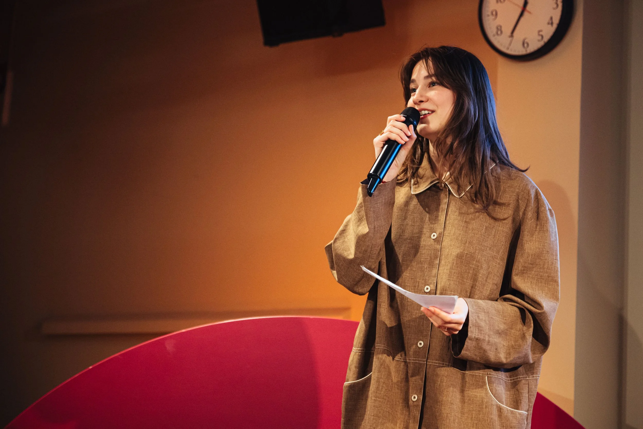 A young woman with long brown hair holding a microphone and a piece of paper, speaking in front of a beige wall with a round clock, wearing a brown oversized button-up shirt.