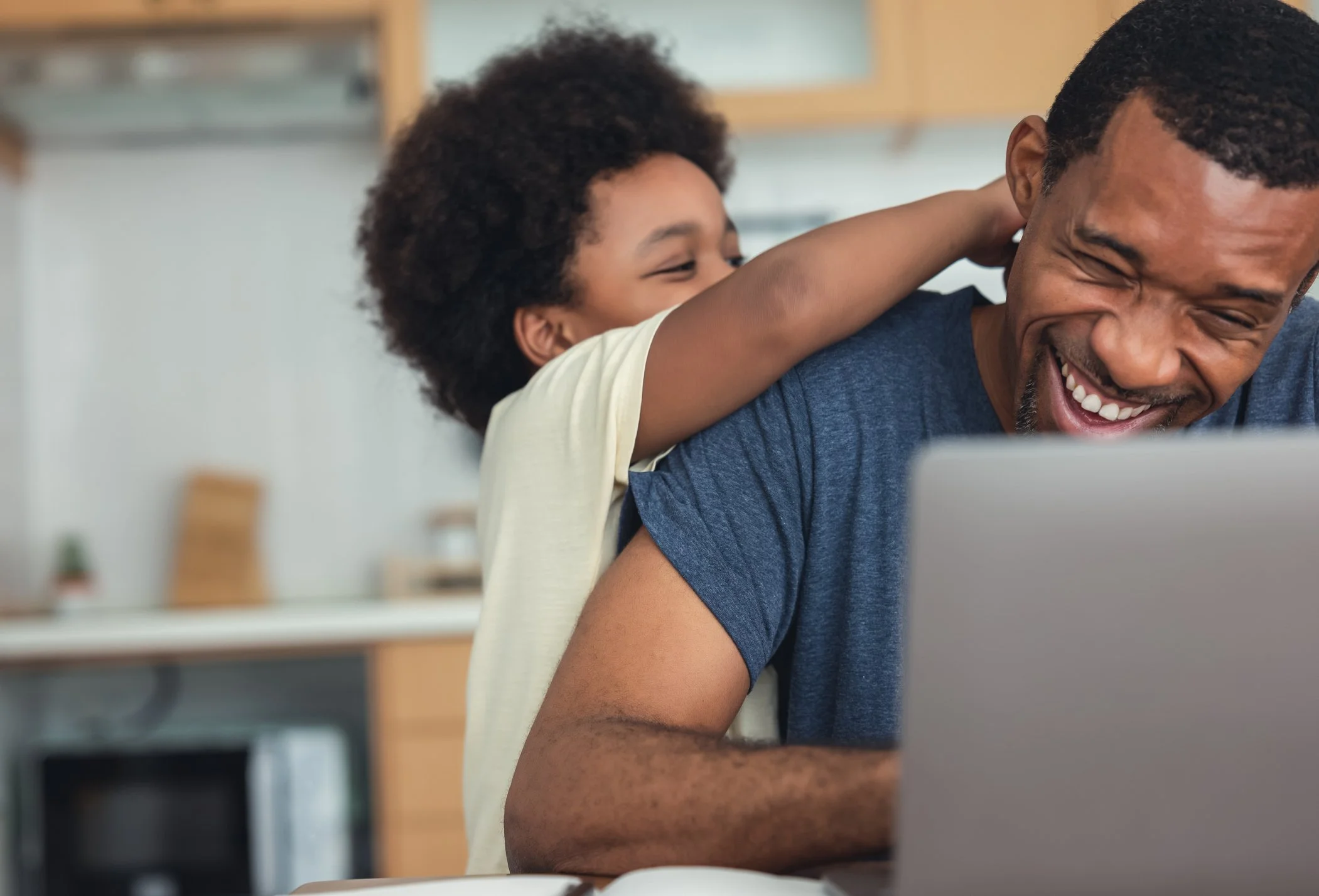 A young girl playfully tugging on her father's ear while sitting at a laptop in a kitchen.