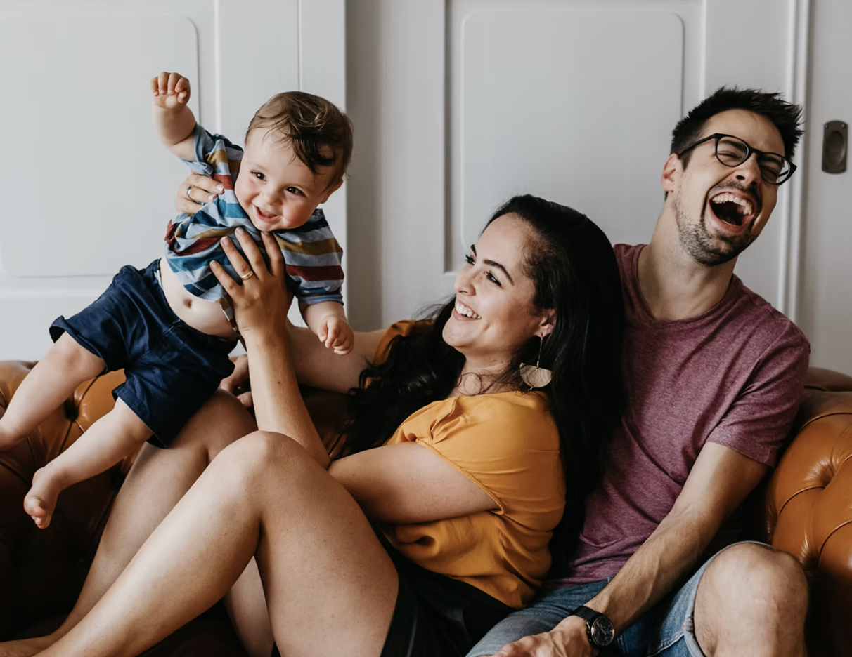Family having fun on a brown leather couch, with a young child being lifted by a woman, all smiling and laughing