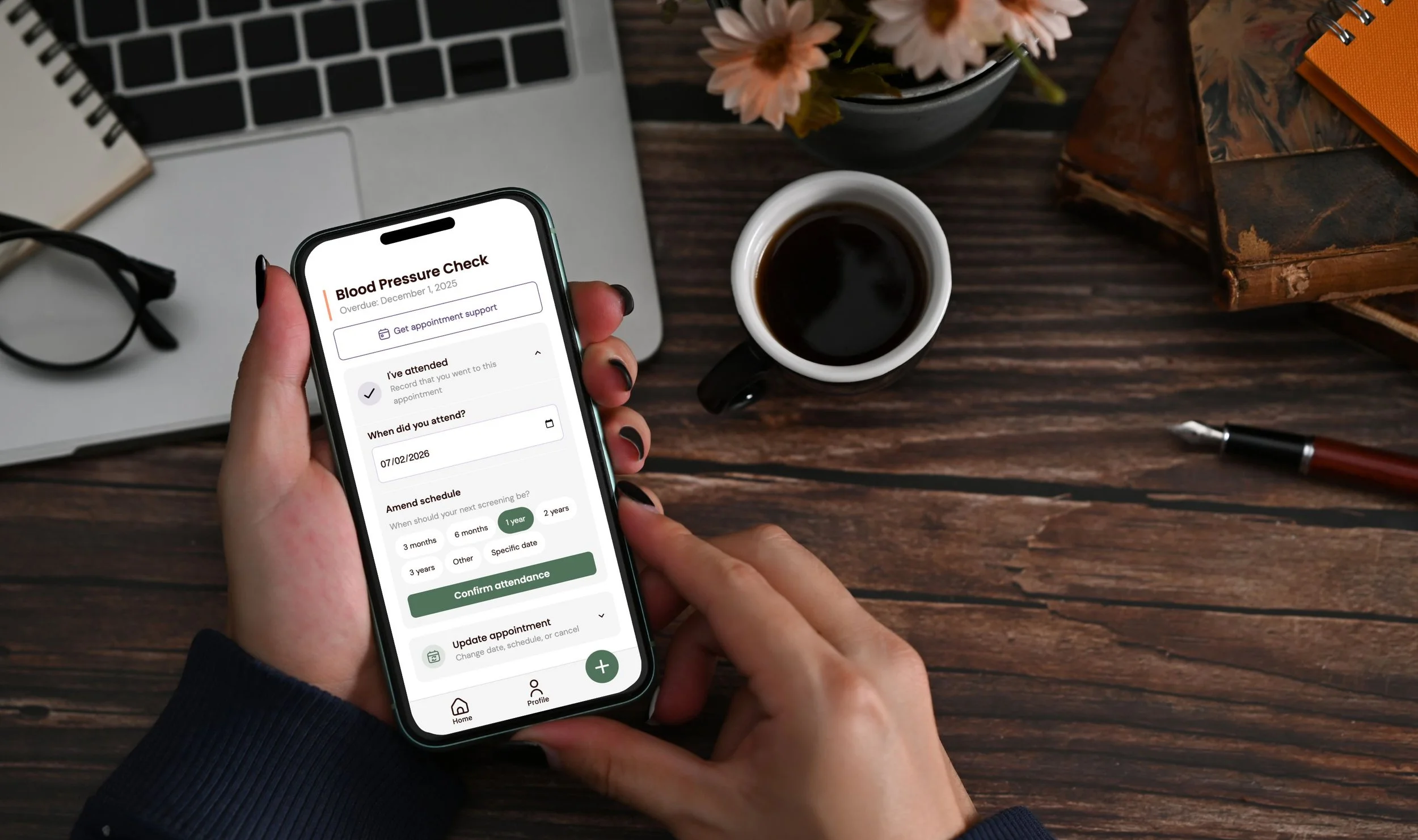 Person holding a smartphone with a blood pressure check app open, sitting at a wooden table with a cup of coffee, a laptop, glasses, a pen, a notebook, and a flowerpot.