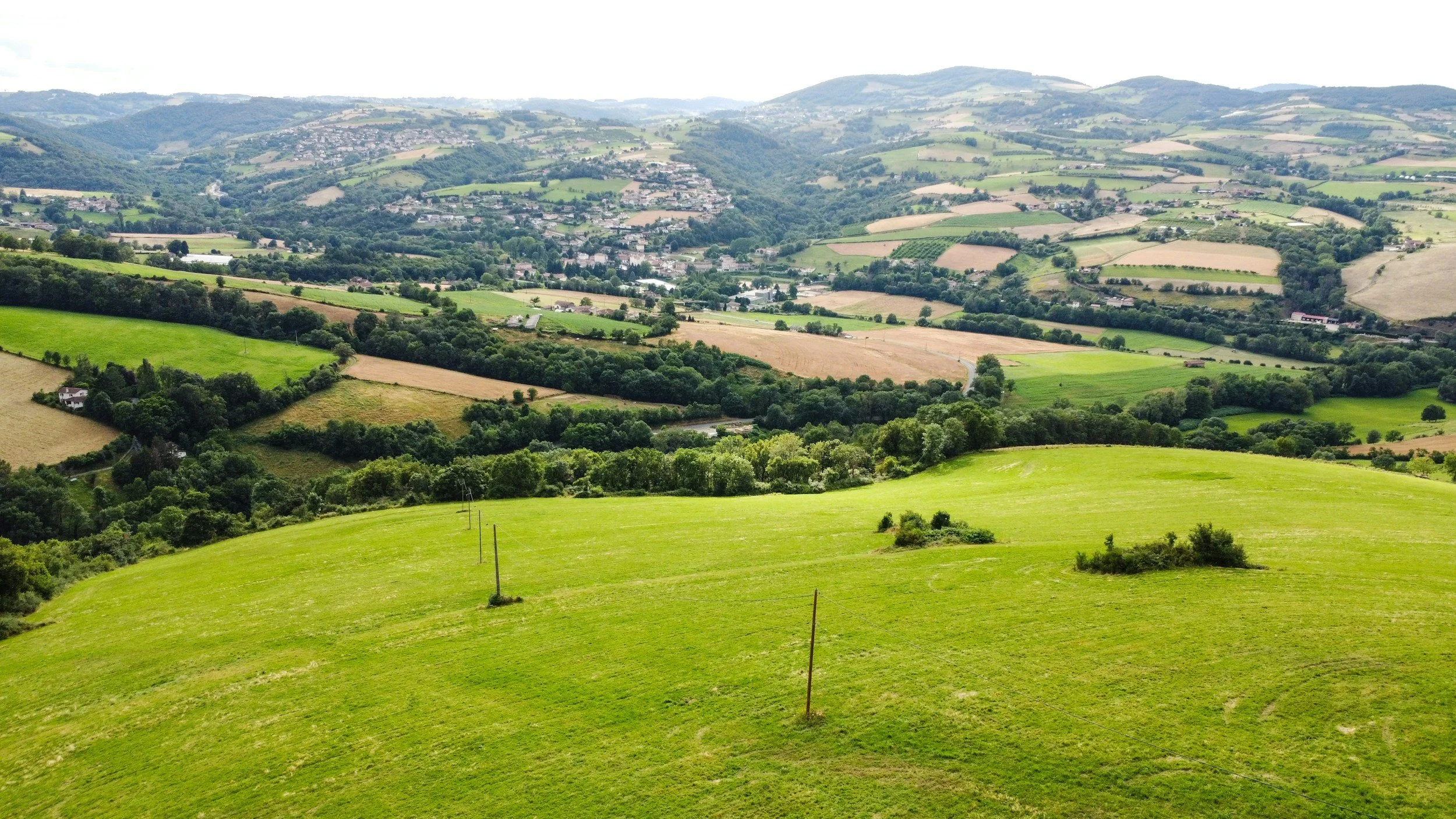 Lush green rolling hills with patchwork farmland and distant town under cloudy sky.