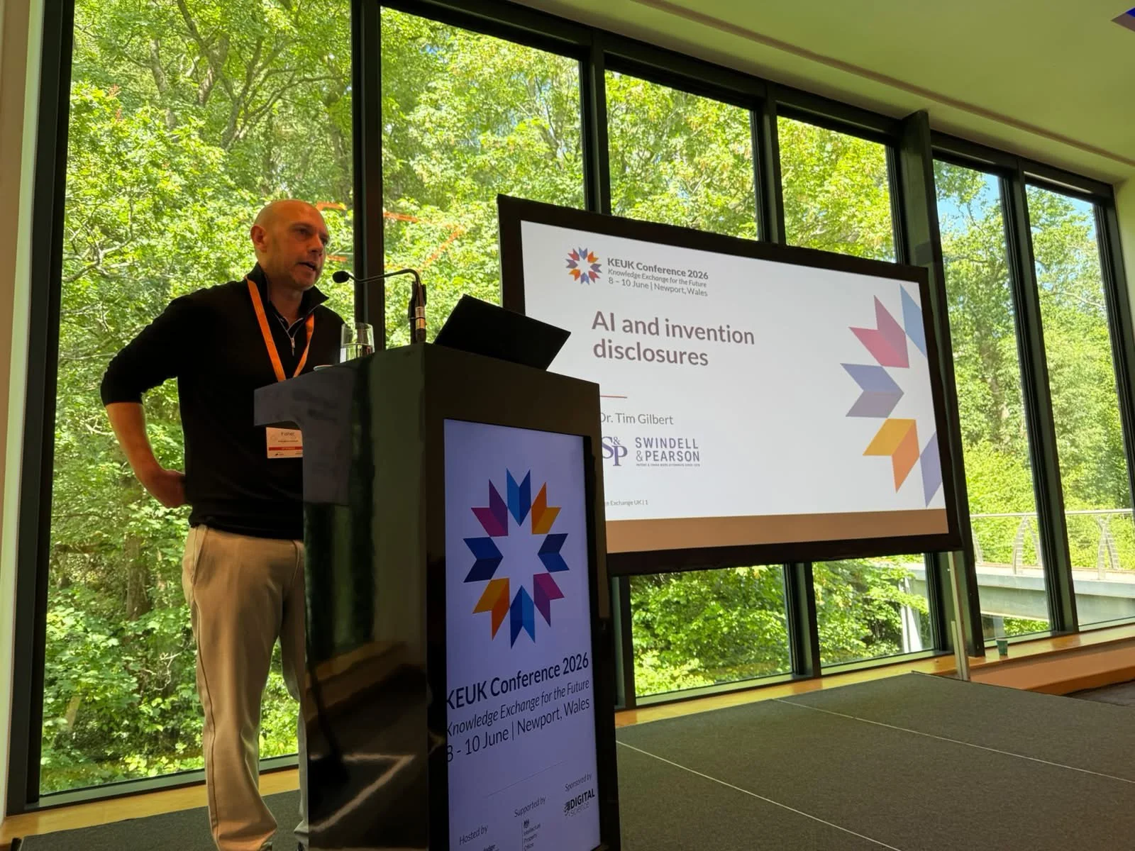 A man with a shaved head and light-colored pants giving a presentation at the KEUK Conference 2026 in Newport, Wales. He is standing at a podium with a large screen behind him displaying the presentation title "AI and invention disclosures" and the conference details.