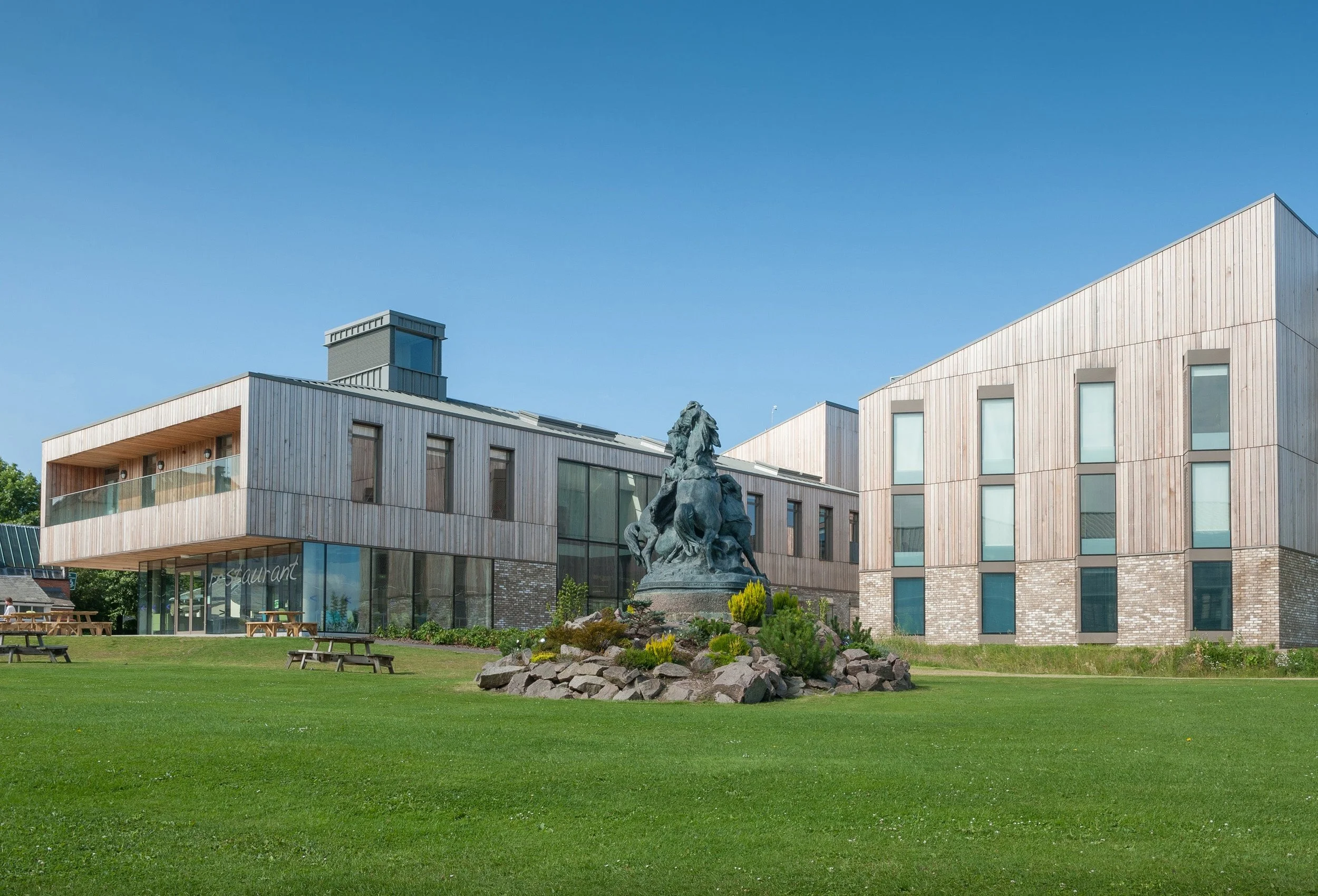 Modern building with wooden siding, large windows, a statue in front, and a grassy area with picnic tables.