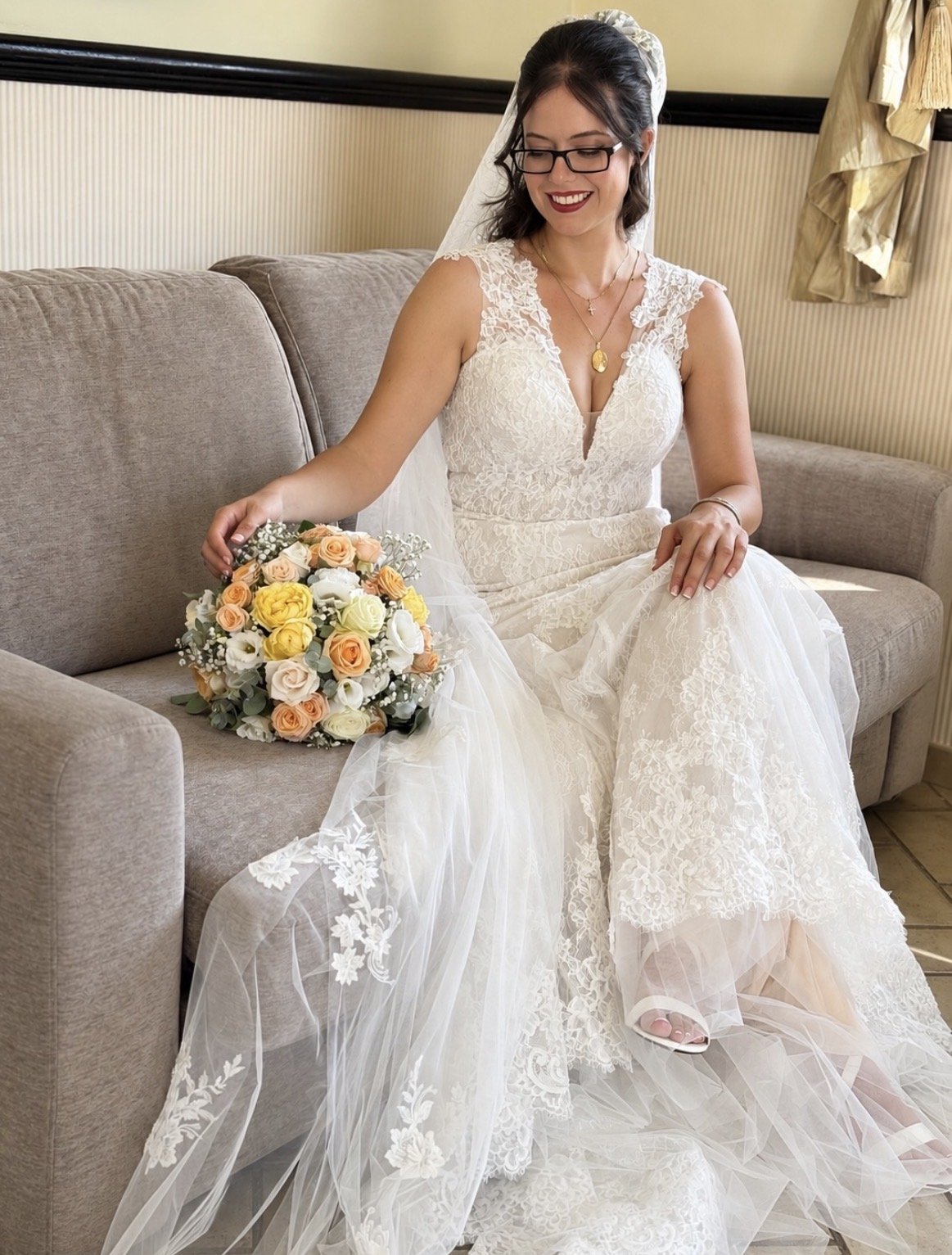A bride in a white lace wedding gown sitting on a beige sofa, holding a bouquet of yellow, peach, and white roses, and wearing glasses and jewelry.