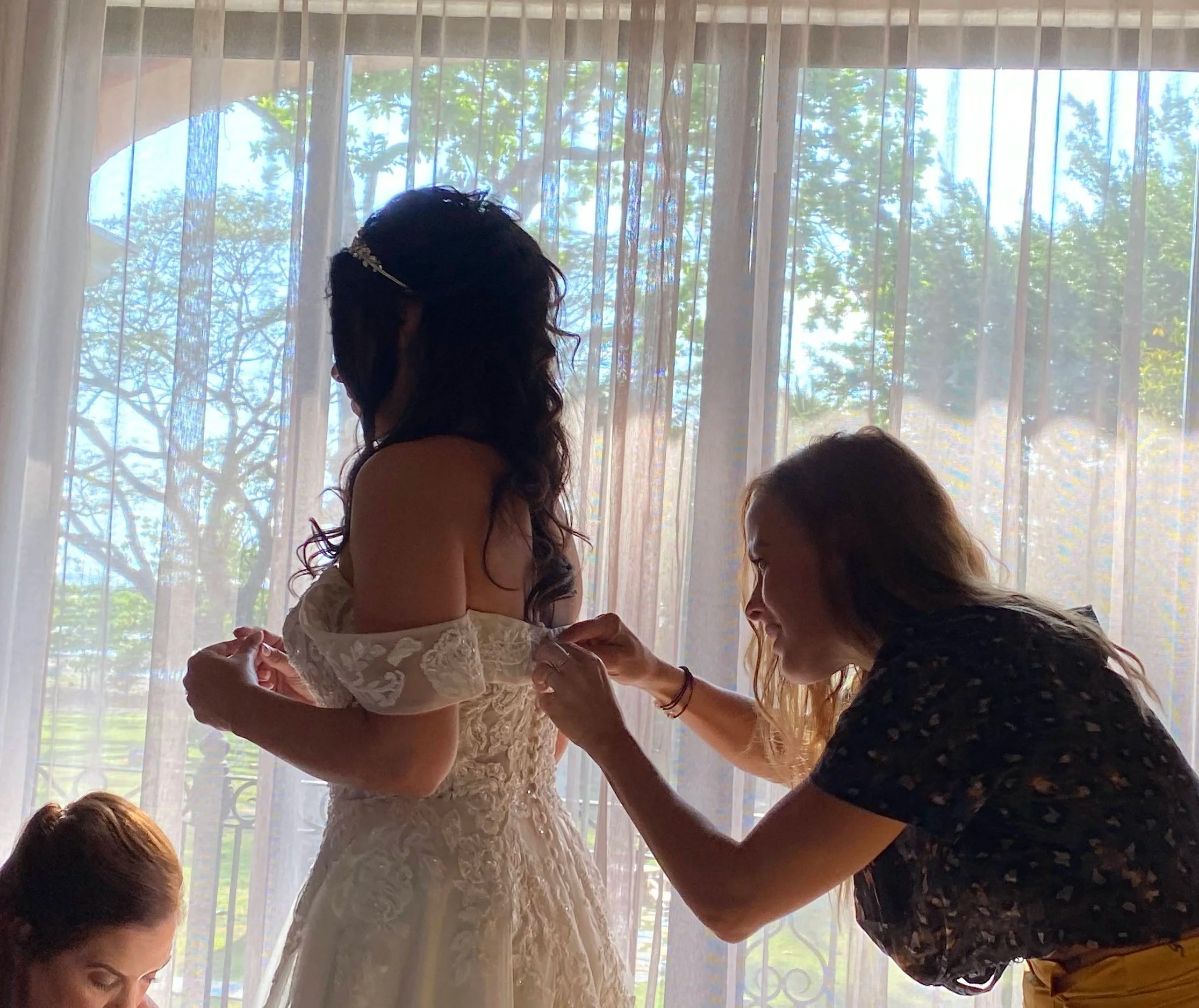 A woman in a wedding dress being helped with the dress by another woman in front of a window with sheer curtains, with trees visible outside. Styled by Cecilia Acquaroli in Rome