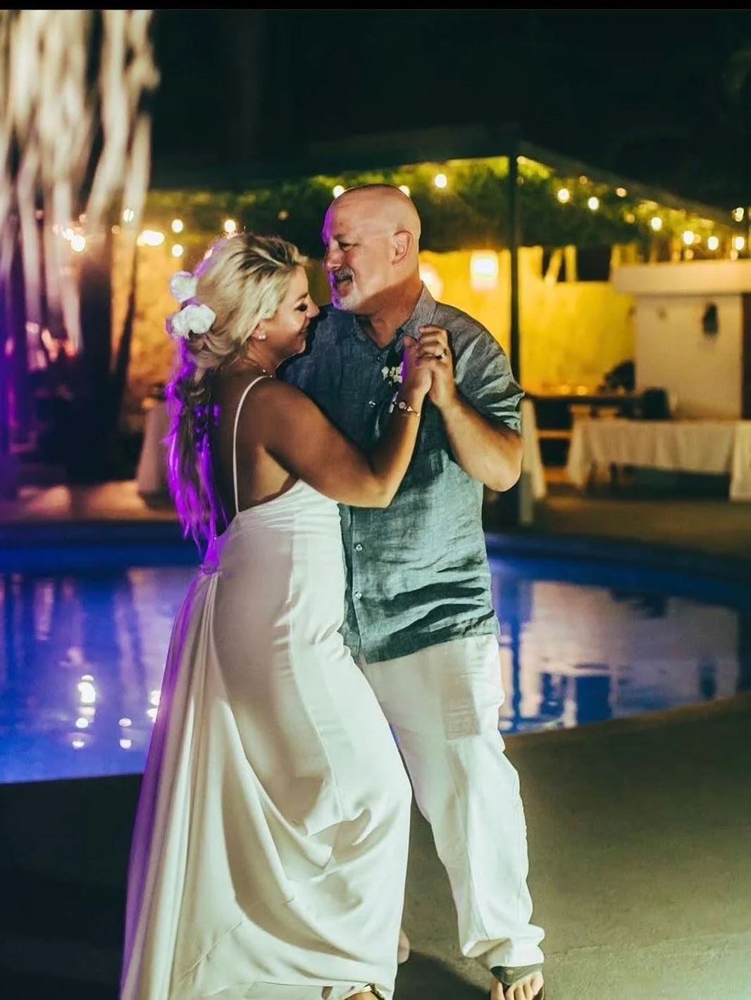 A couple dances by a pool at night, with string lights and a yellow-lit building in the background.