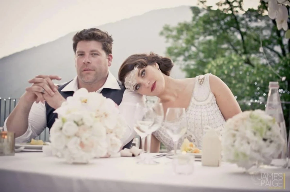 A man and a woman sitting at a decorated outdoor wedding reception table, with the woman resting her head on the man's shoulder. They are surrounded by floral centerpieces and glassware, with a scenic background of trees and mountains.