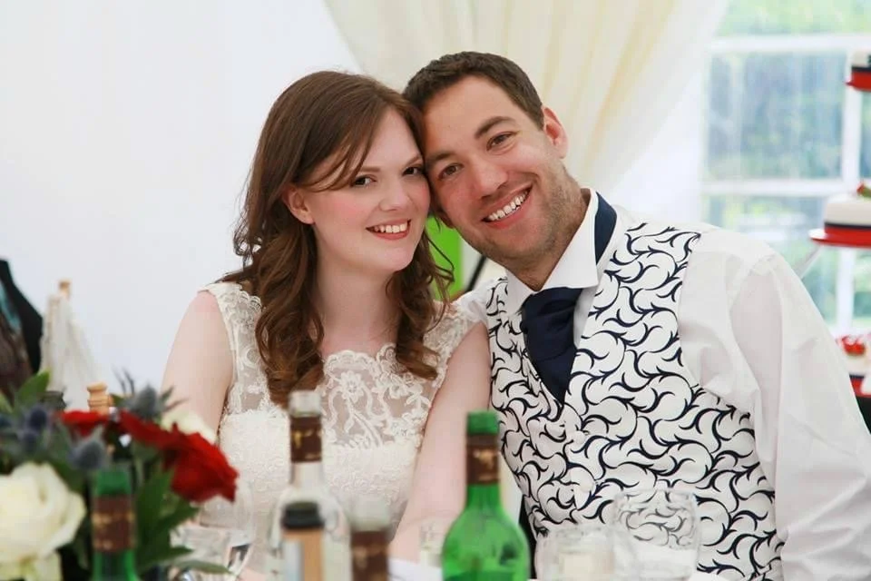 Happy couple at a wedding reception, smiling and close together, with bottles and flowers in the foreground.