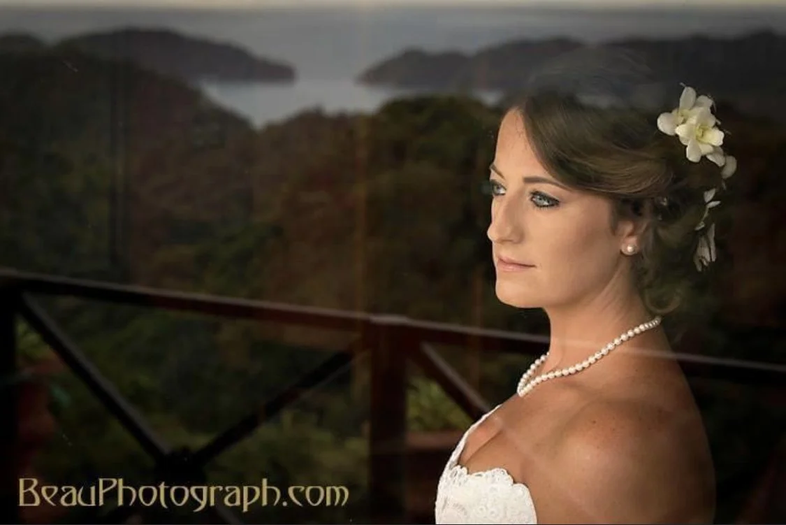 A bride with light skin and blonde hair adorned with white flowers, wearing pearl jewelry and a strapless white dress, looking thoughtfully to the side with a scenic outdoor background. Styled by Ceciia Acquaroli in Rome